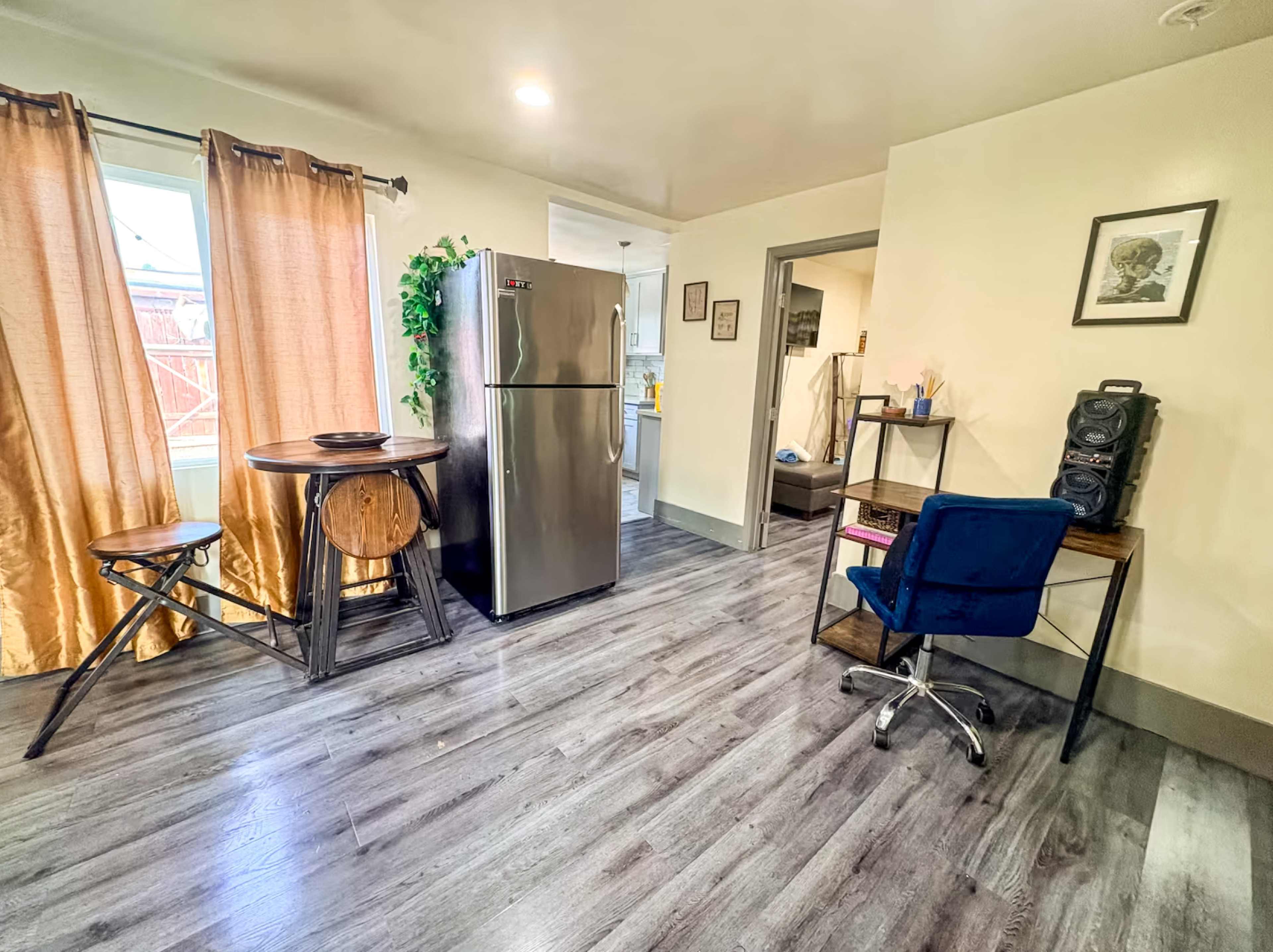 The image shows a small room featuring a kitchen area with a refrigerator, a round wooden table, a desk with a blue chair, and large curtains covering a window.