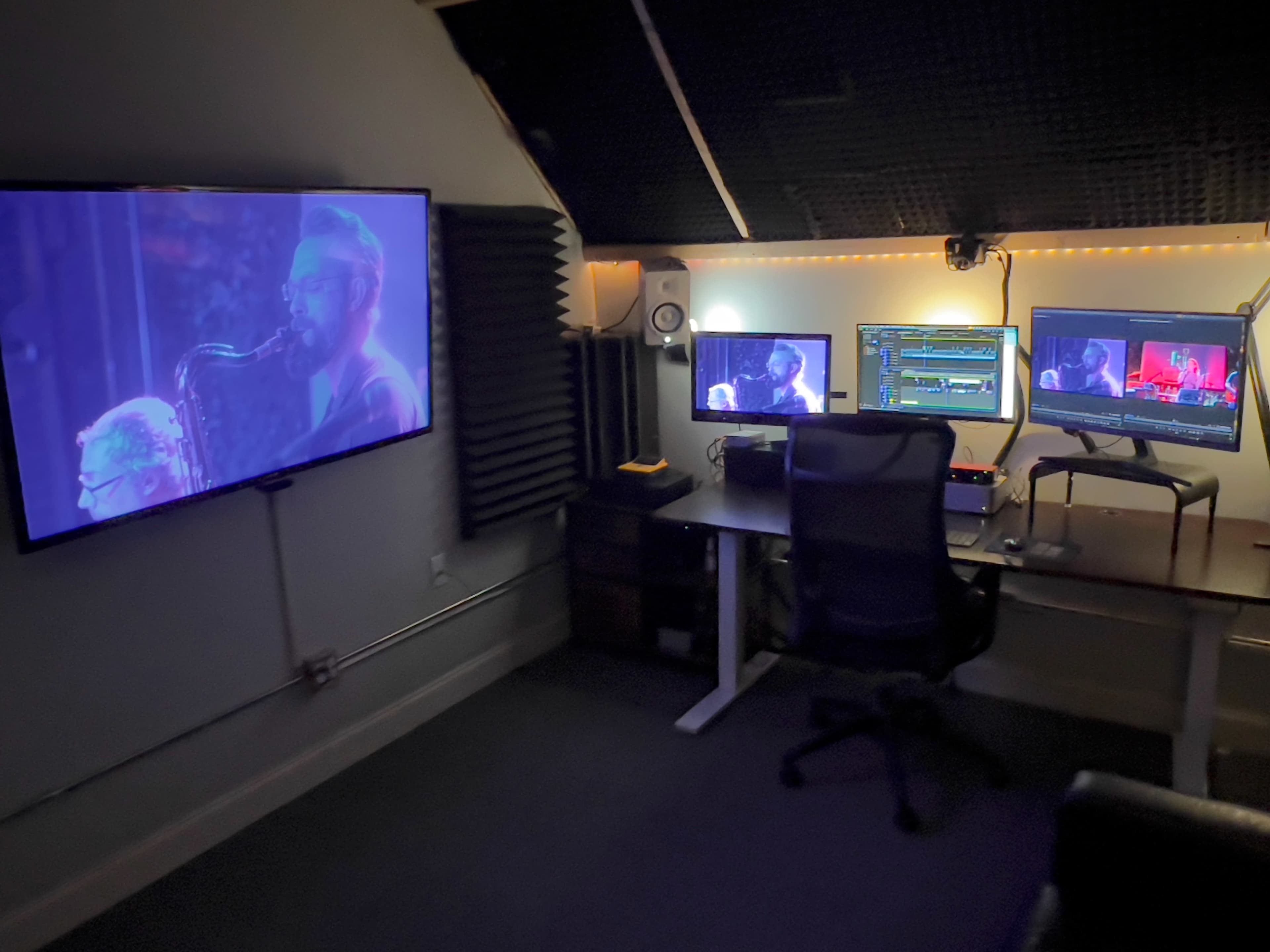 The image shows a dimly lit recording studio with multiple screens displaying a live performance and sound equipment.