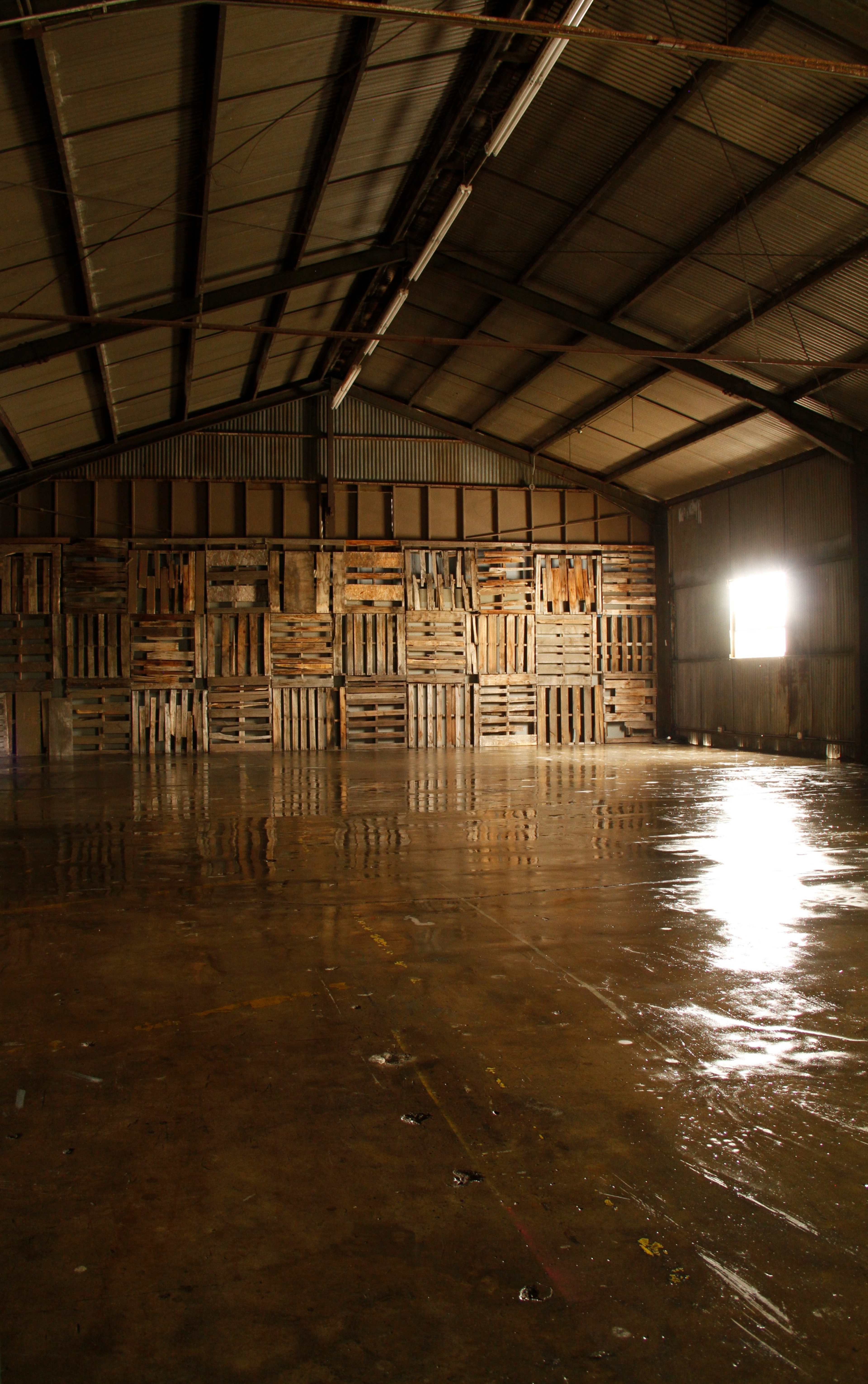 The image shows an empty warehouse interior with wooden pallets stacked against a back wall and light streaming in from a window.