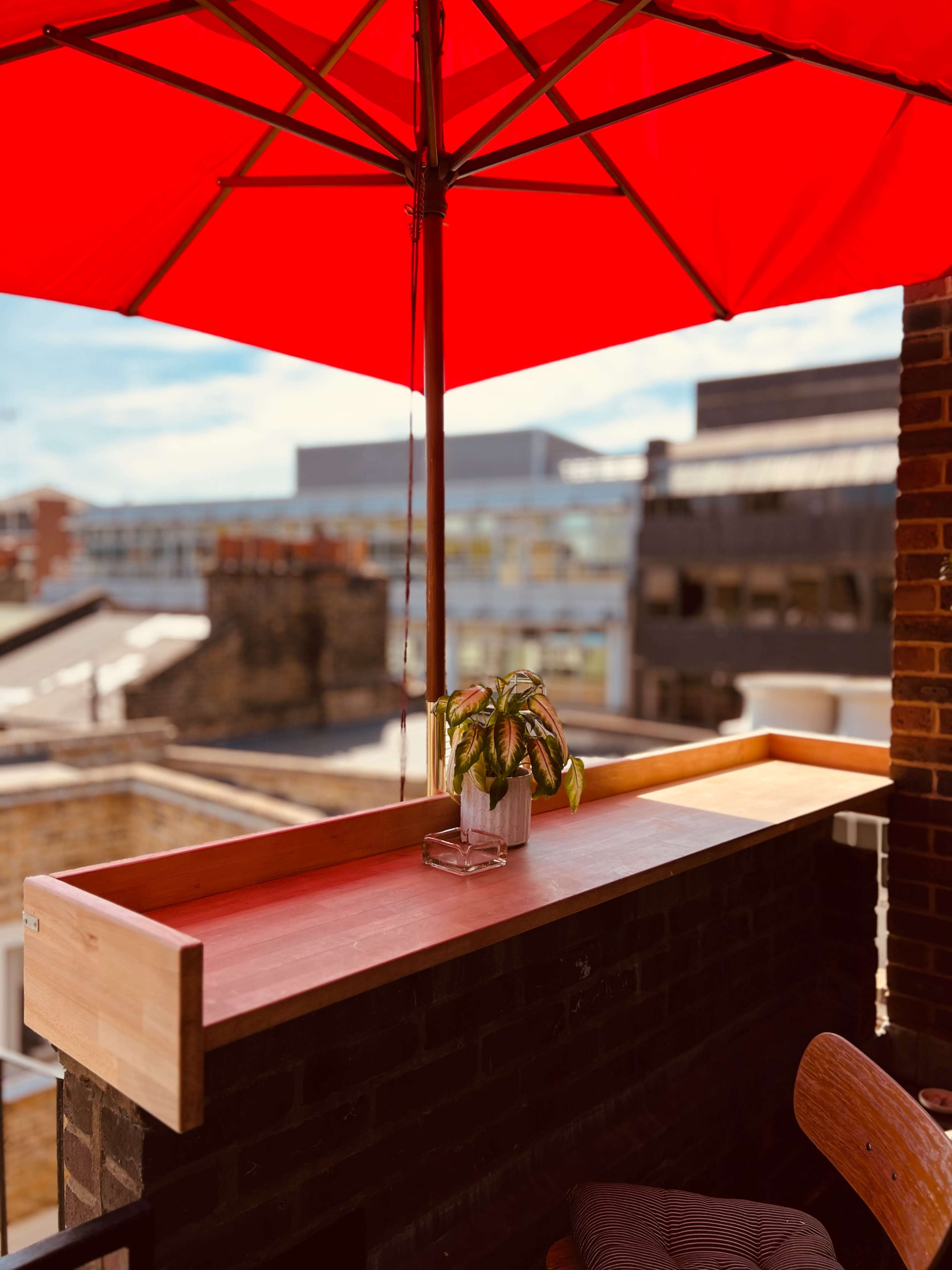 A bright red umbrella shades a wooden ledge holding a small potted plant on a balcony overlooking city buildings.