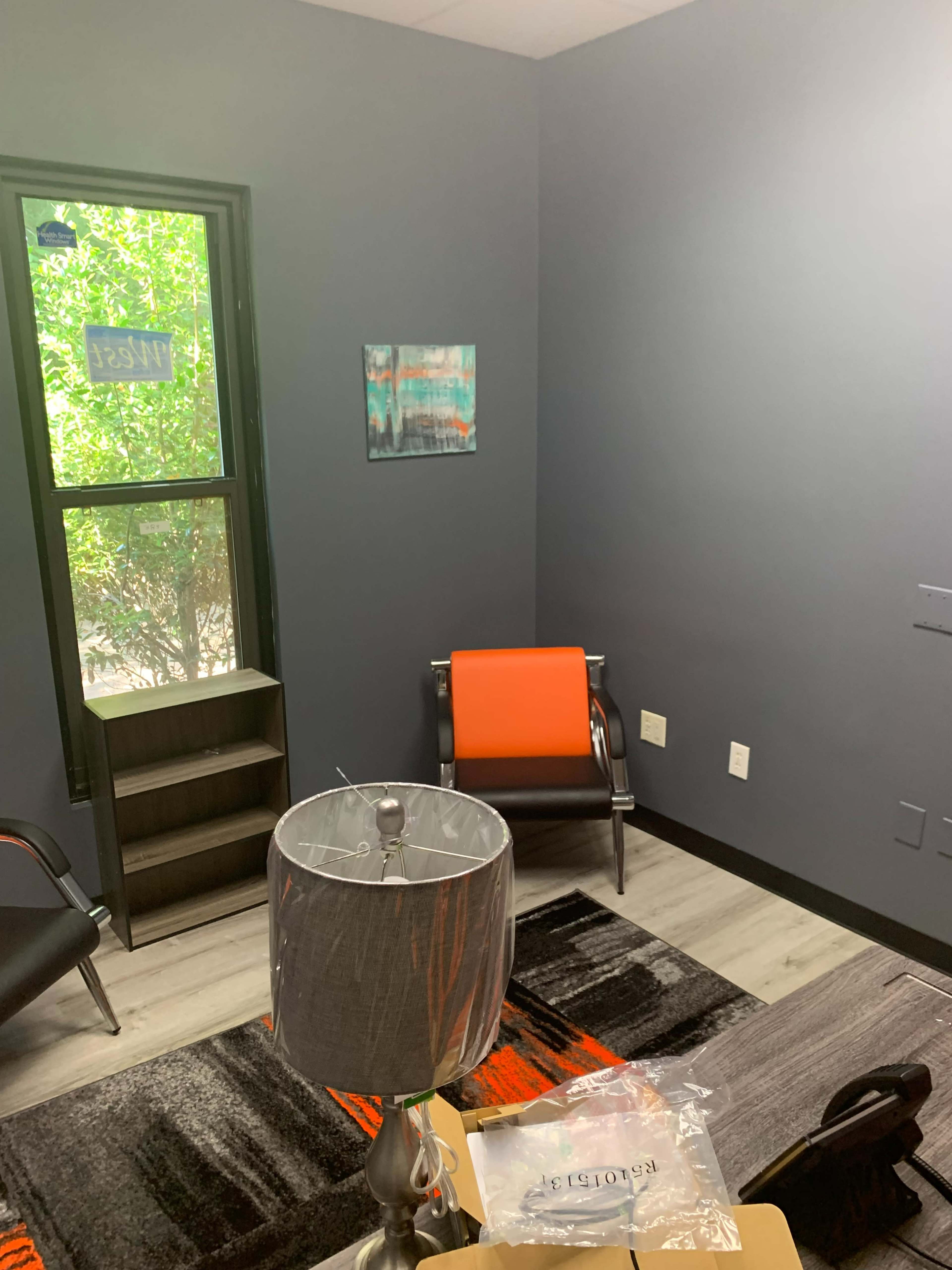 A small office space with gray walls, a window, a colorful painting, a lamp, and orange-accented furniture.