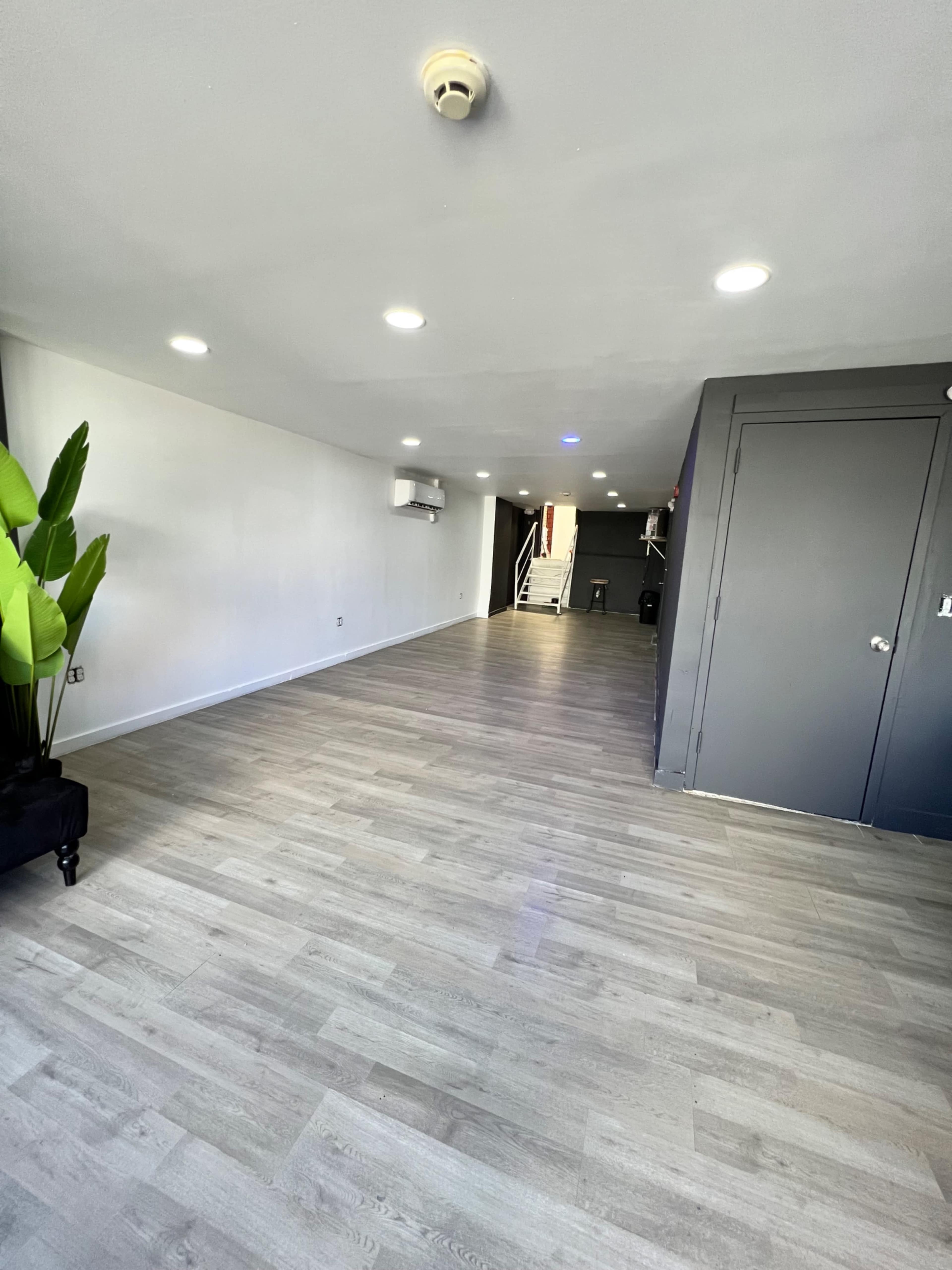 A spacious, empty room with light-colored wood flooring, white walls, and a staircase in the background.
