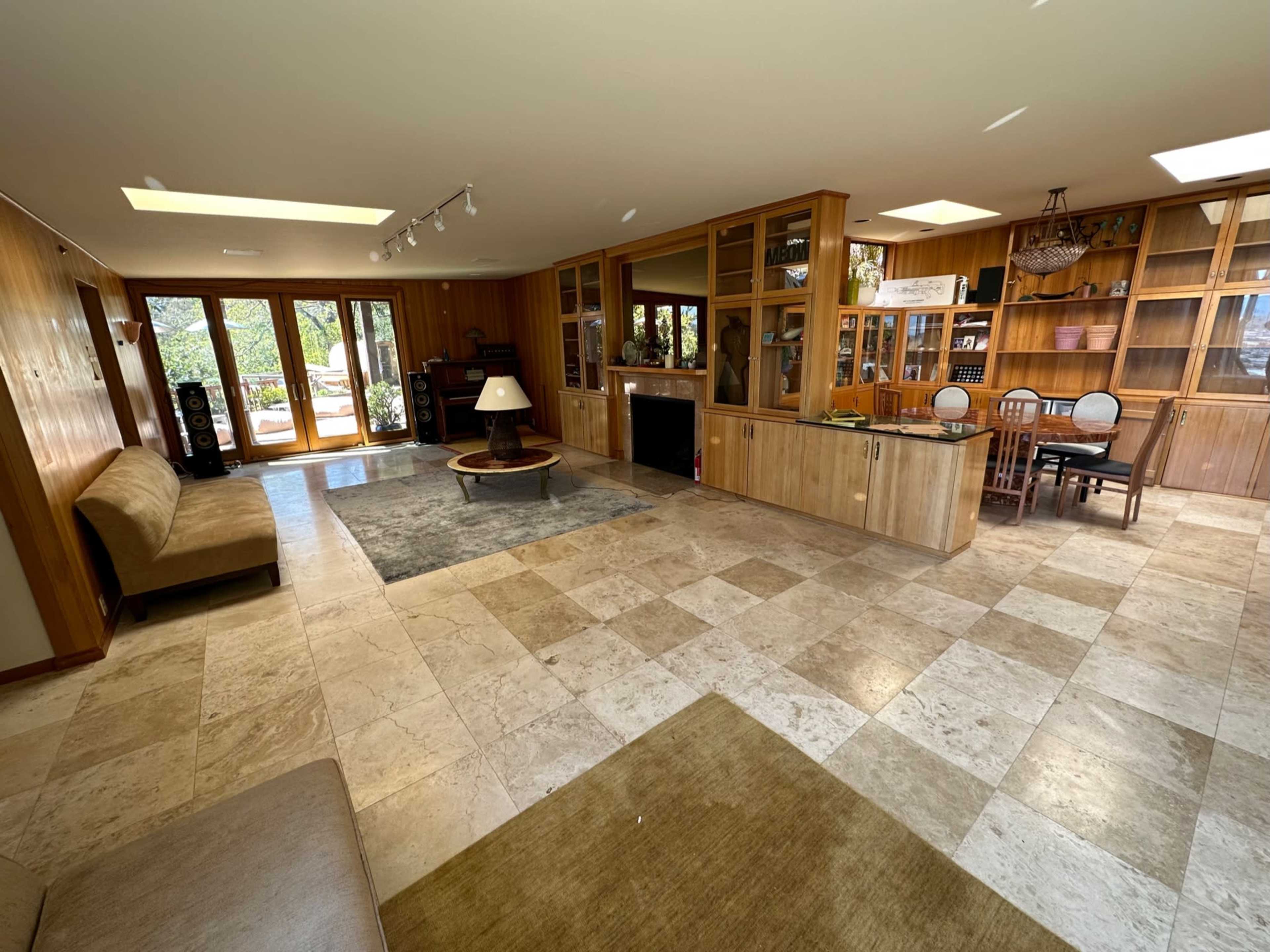 The image depicts a spacious room with tiled floors, wooden paneling, a seating area, a fireplace, and built-in shelves, with large windows that open to an outdoor view.
