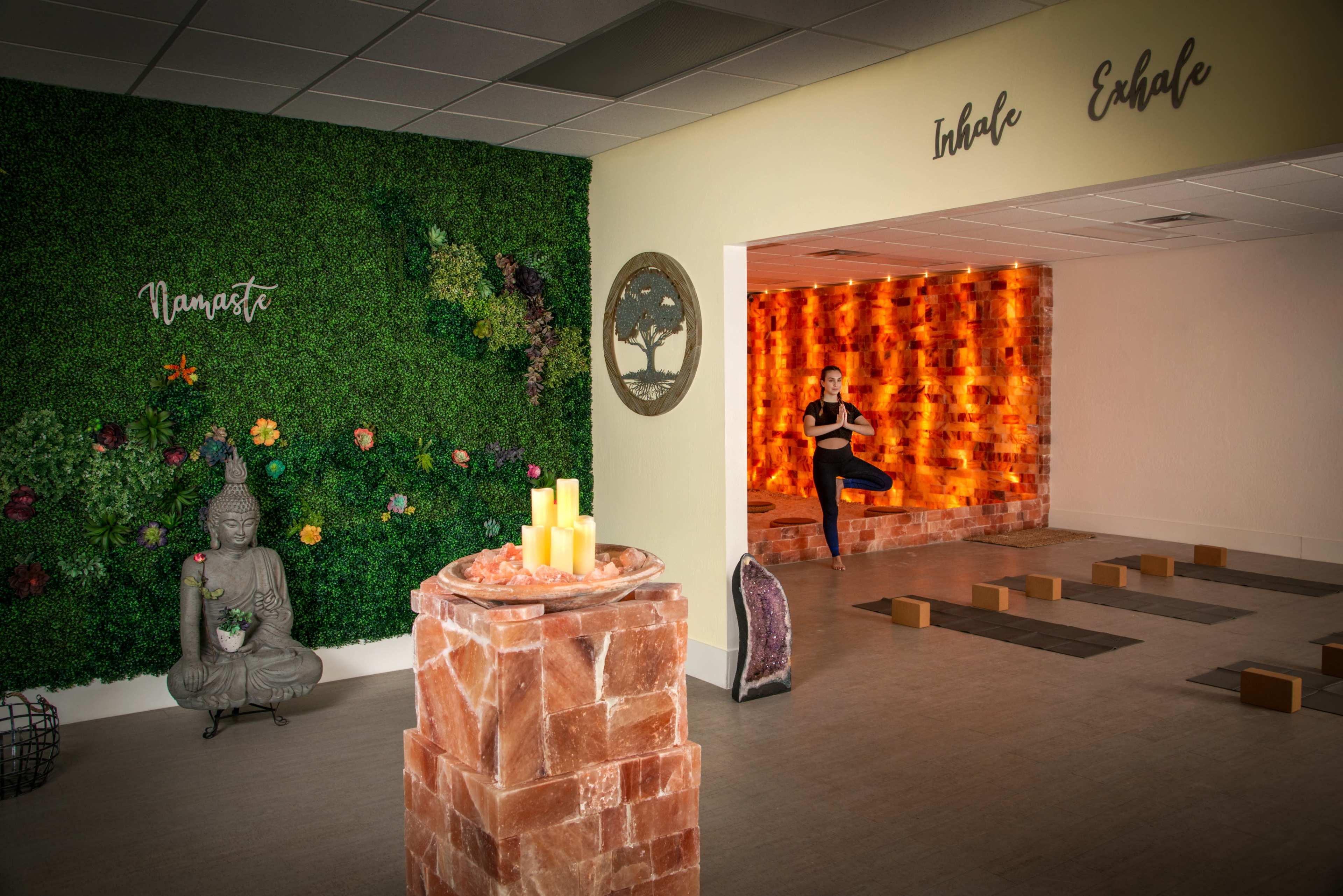 A serene wellness space with a salt block wall, a meditation area featuring a Buddha statue, and a woman performing a yoga pose in the background.