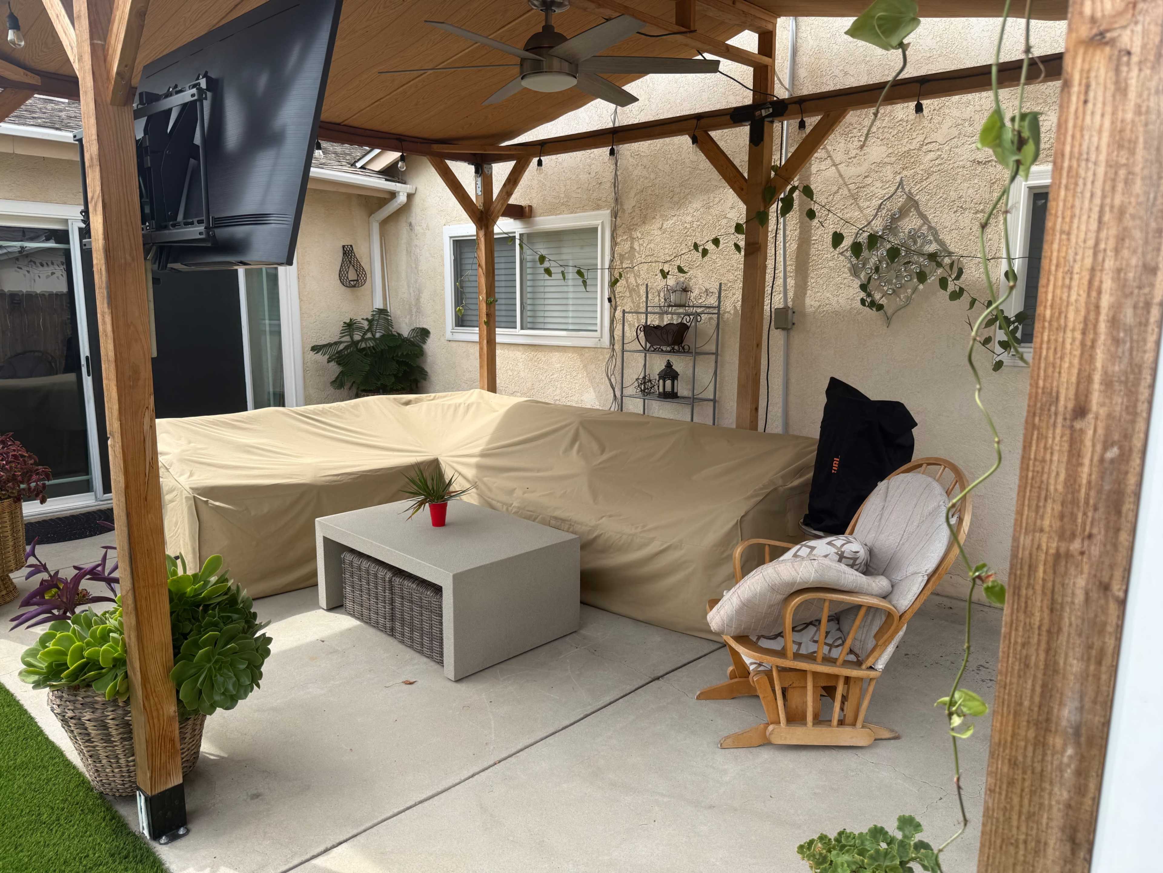 The image shows a covered patio area with a large beige sectional sofa, a small coffee table, a rocking chair, and various potted plants.