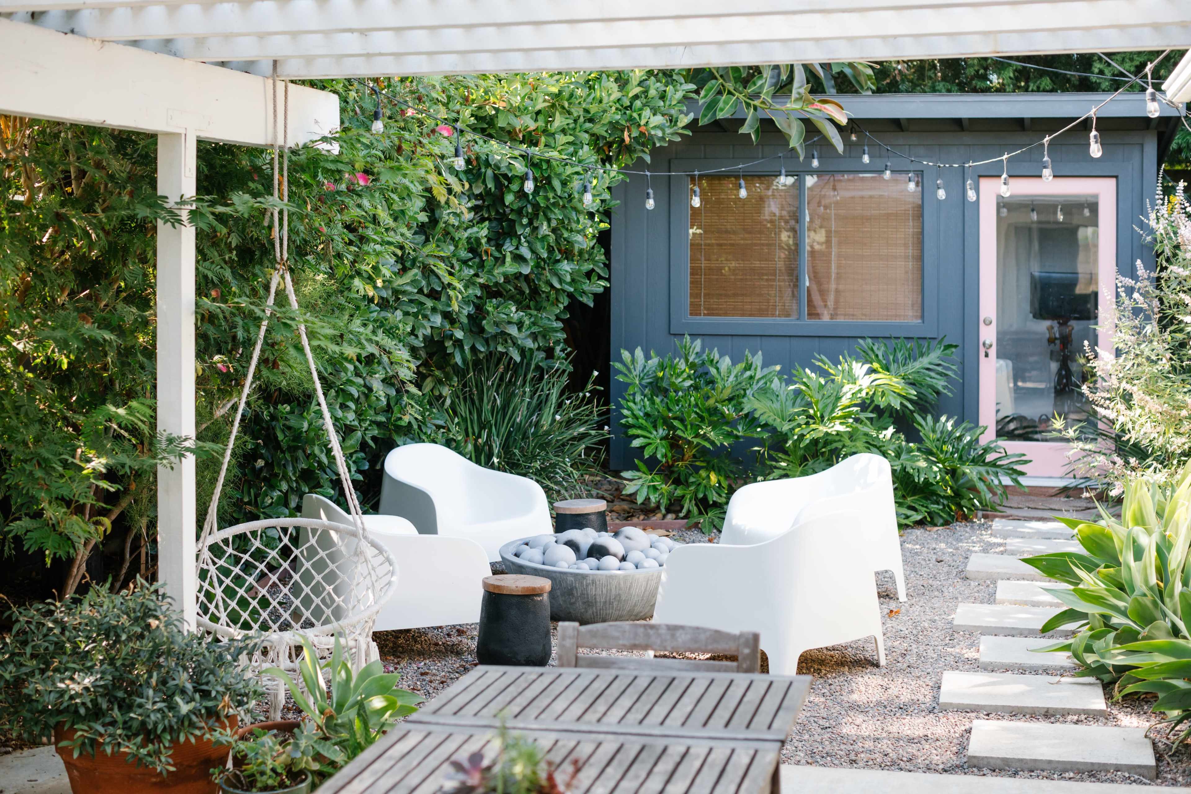 A cozy outdoor space features white chairs surrounding a stone fire pit, with a small, dark-colored shed and lush greenery in the background.