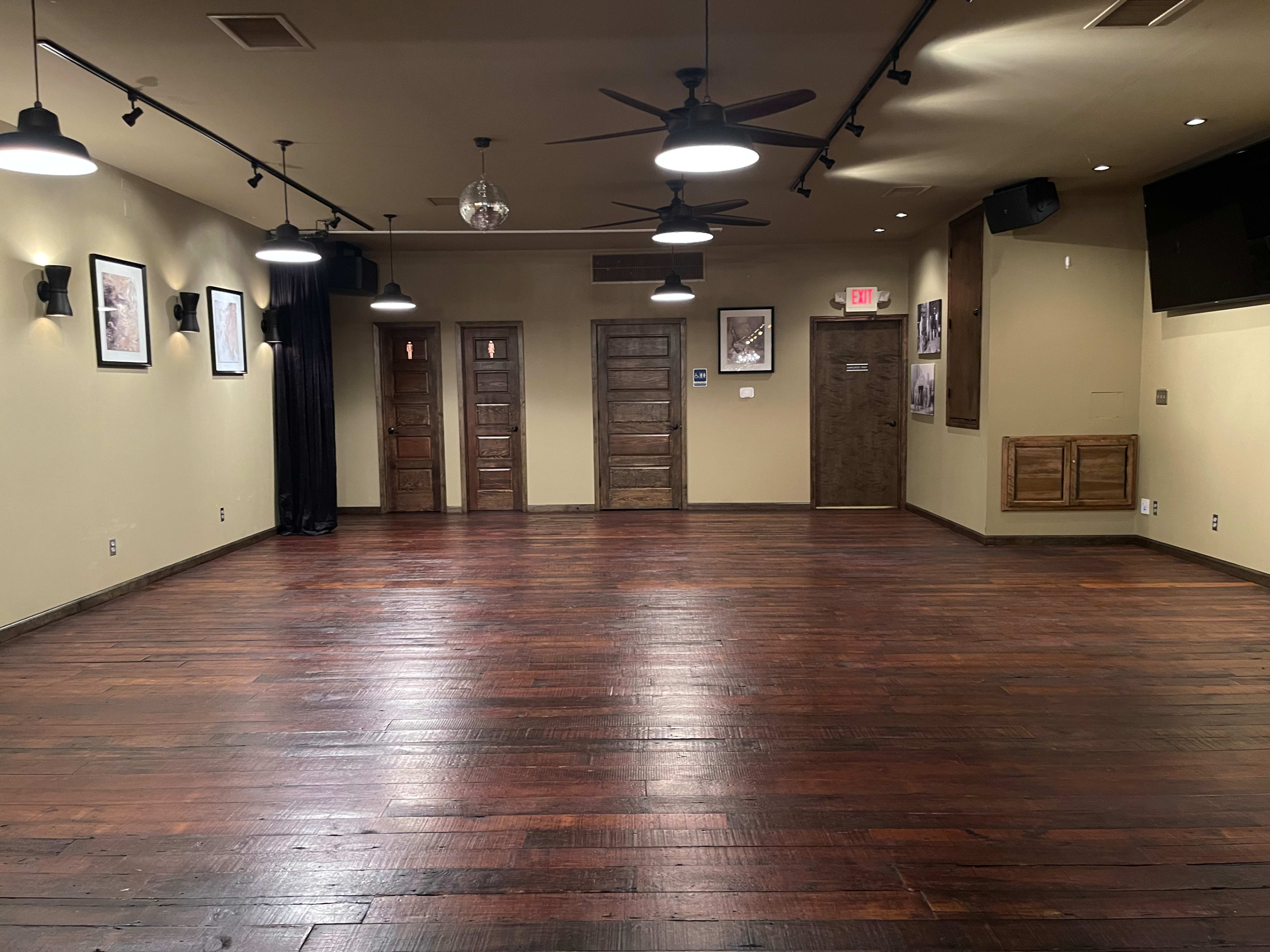 The image shows an empty dance studio with wooden floors, wall art, and three wooden doors.