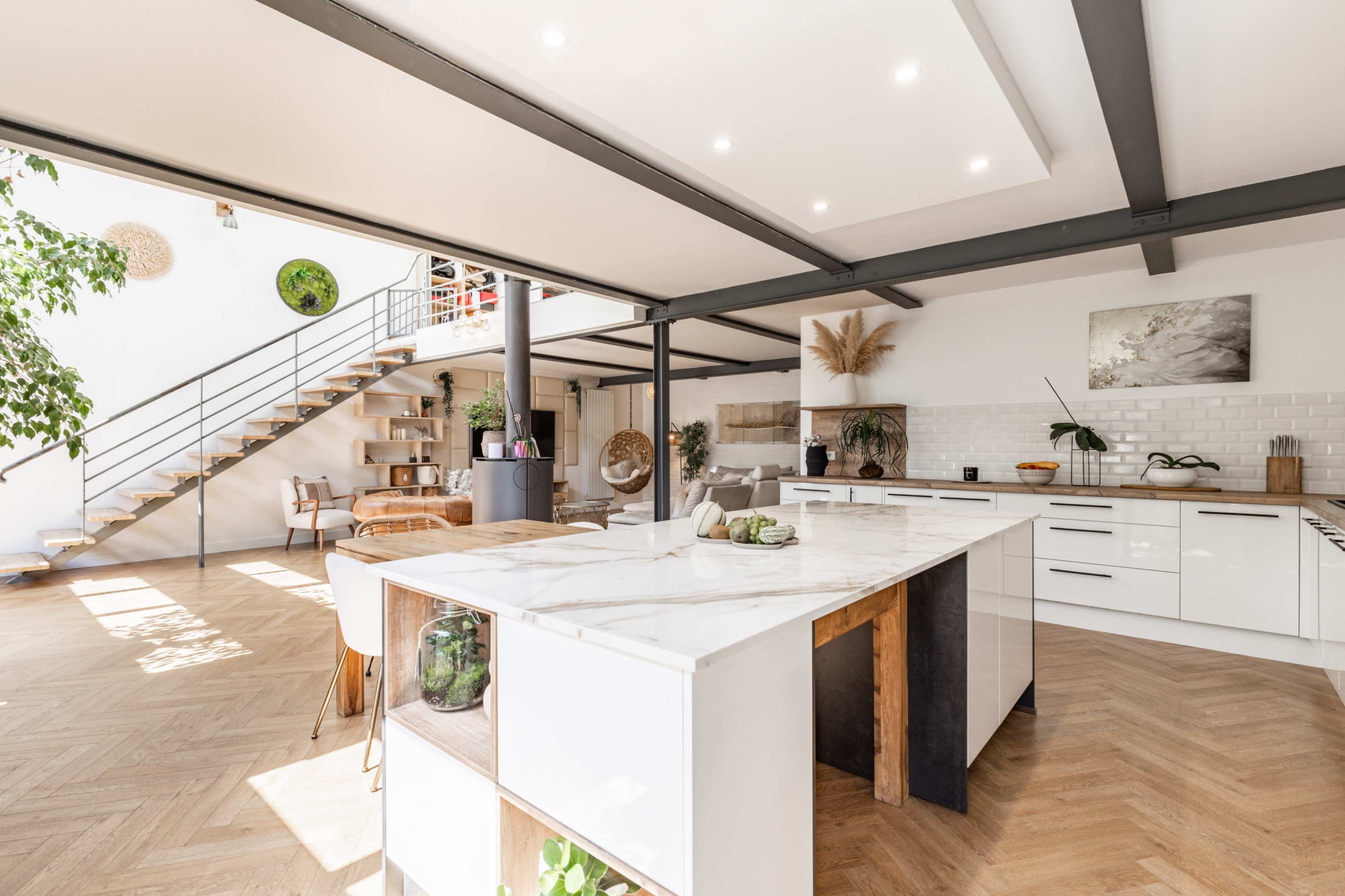 A modern, open-concept kitchen and dining area featuring a large marble island, sleek cabinetry, and a staircase leading to an upper level.