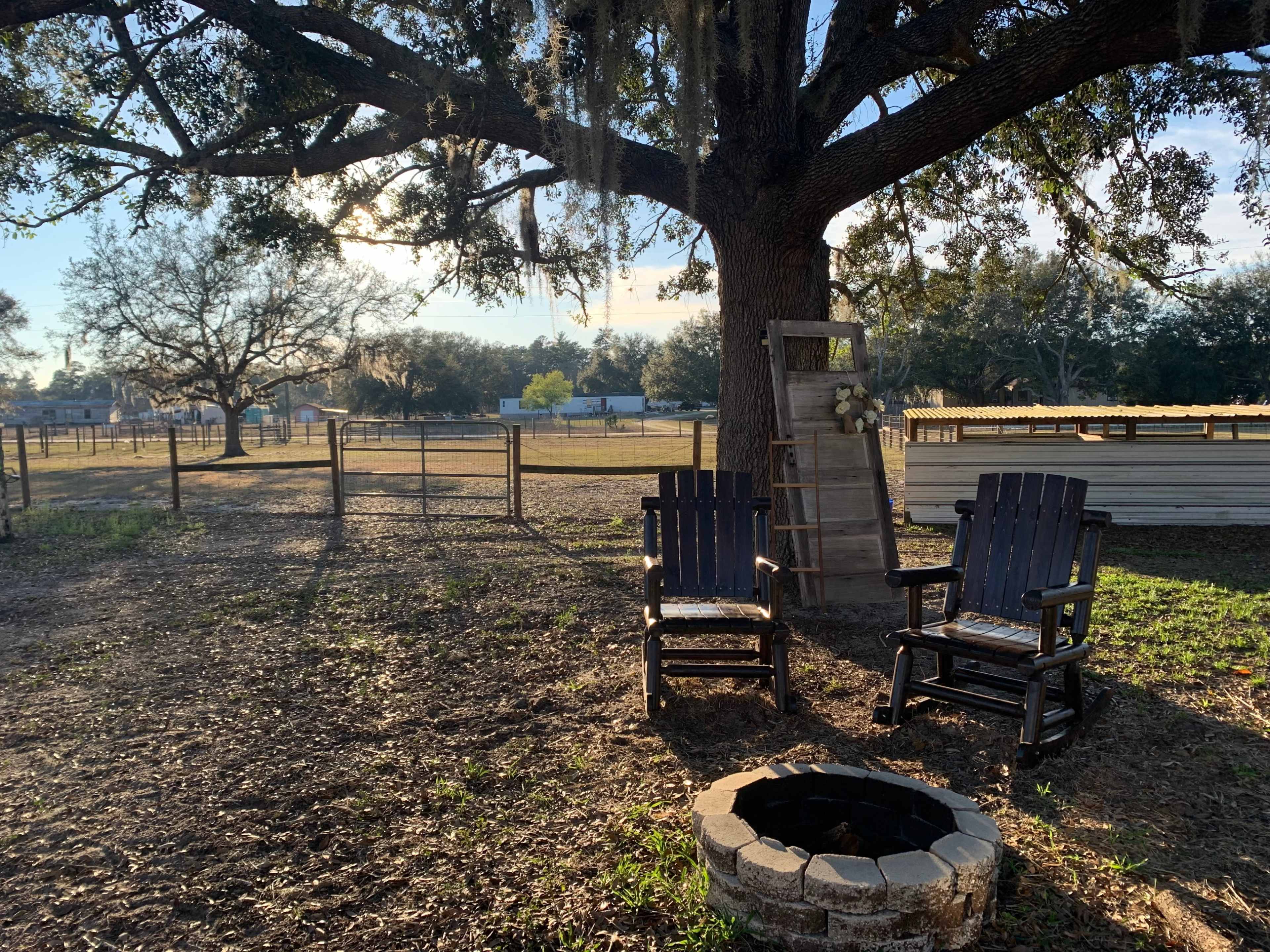 Two wooden rocking chairs sit beside a stone fire pit under a large oak tree in an open field.