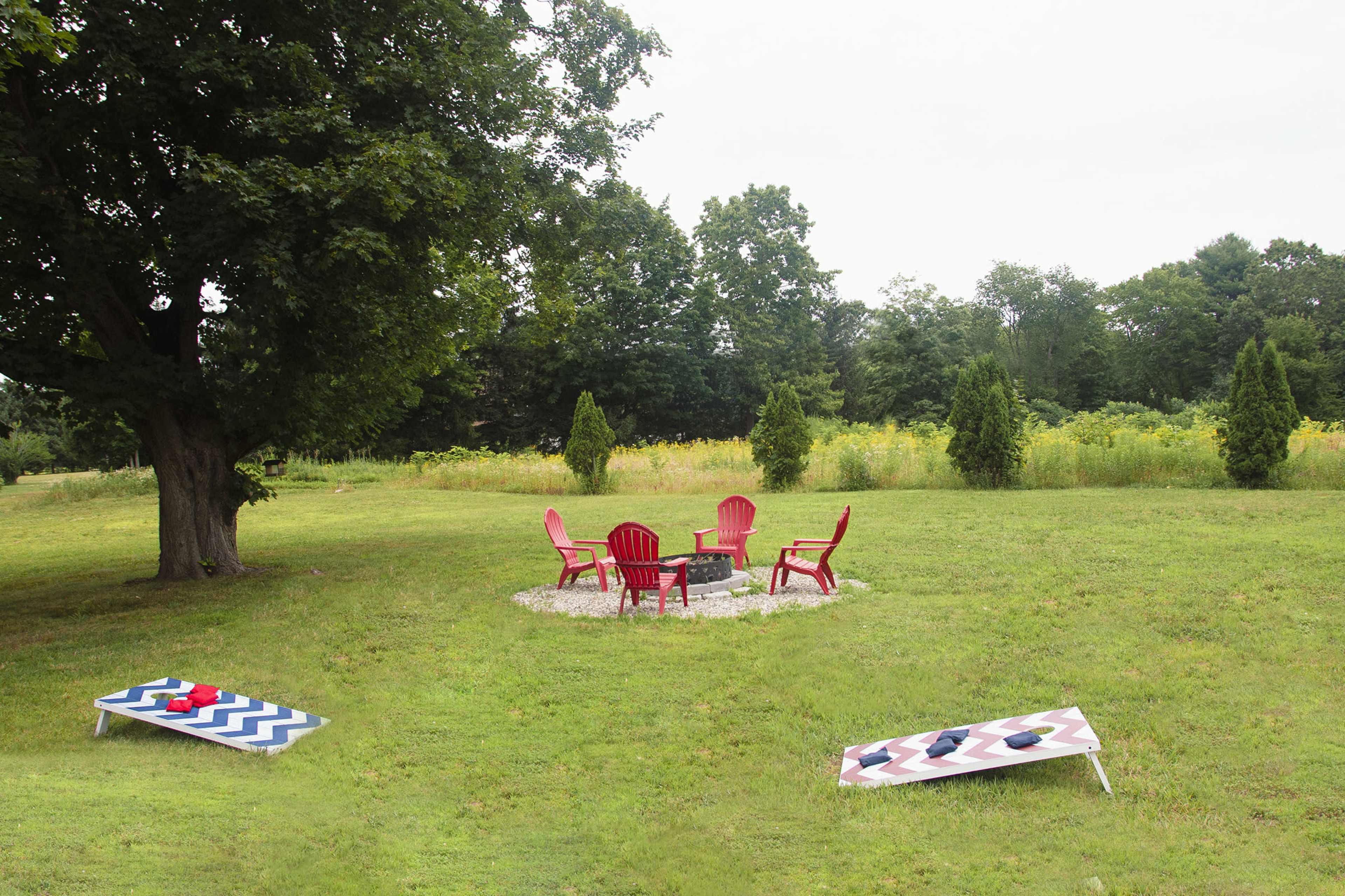 A grassy yard with a circular seating area surrounded by four red chairs and a fire pit, flanked by two bean bag toss boards.