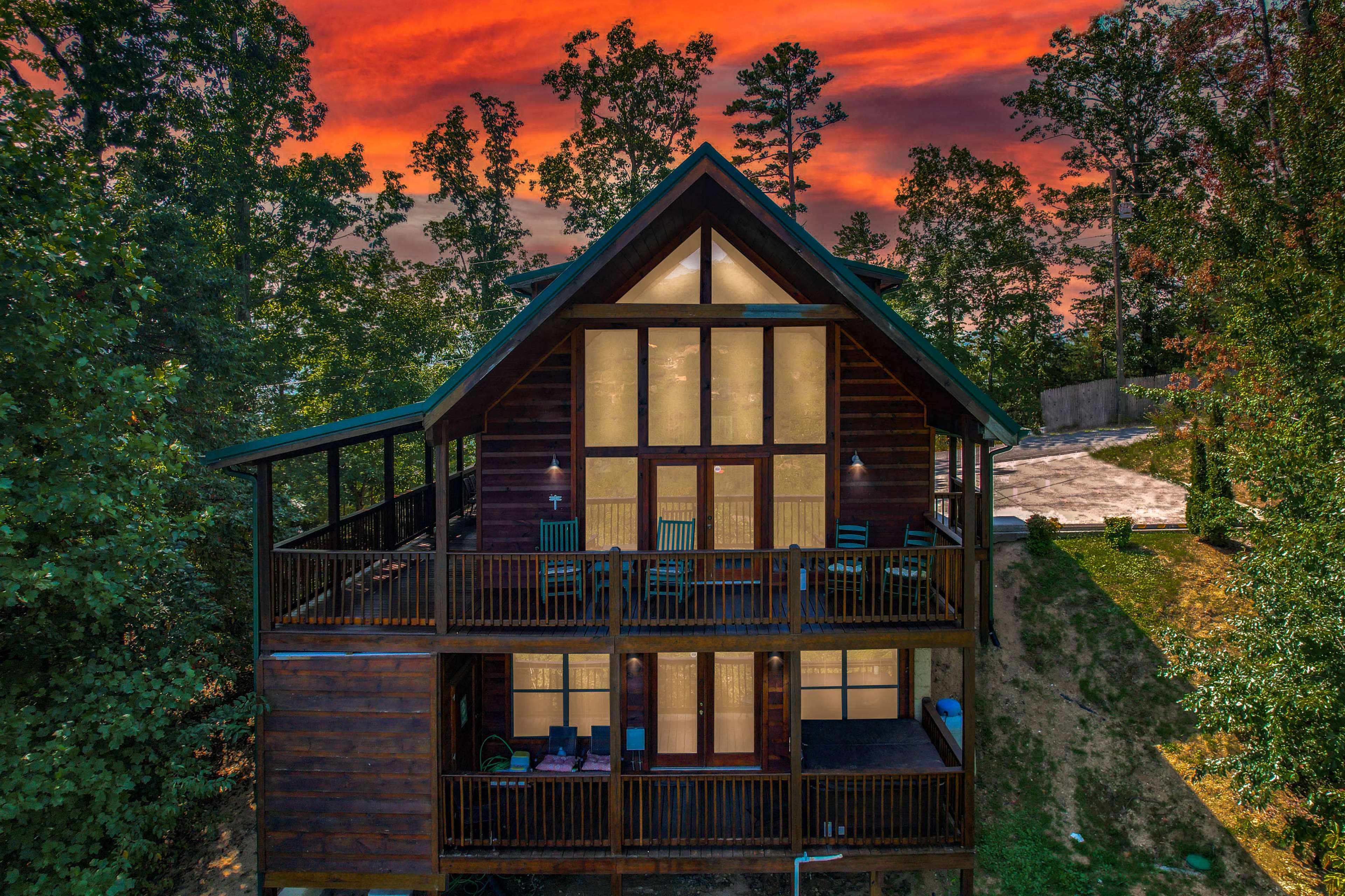 A wooden cabin with large glass windows and multiple balconies is set against a backdrop of a vibrant sunset and surrounded by trees.