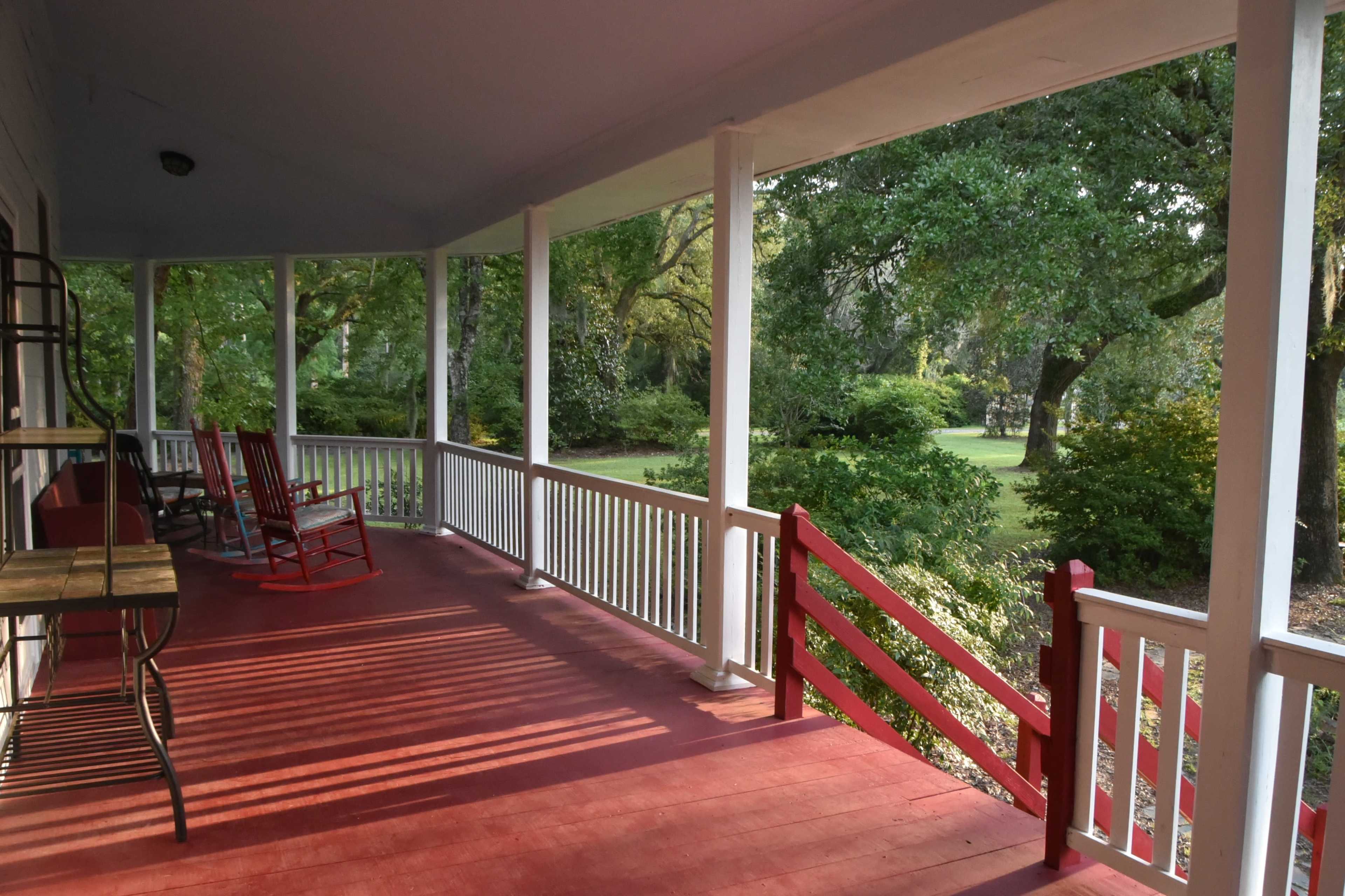 The image shows a spacious porch with red flooring, white columns, and rocking chairs, surrounded by a landscaped garden.