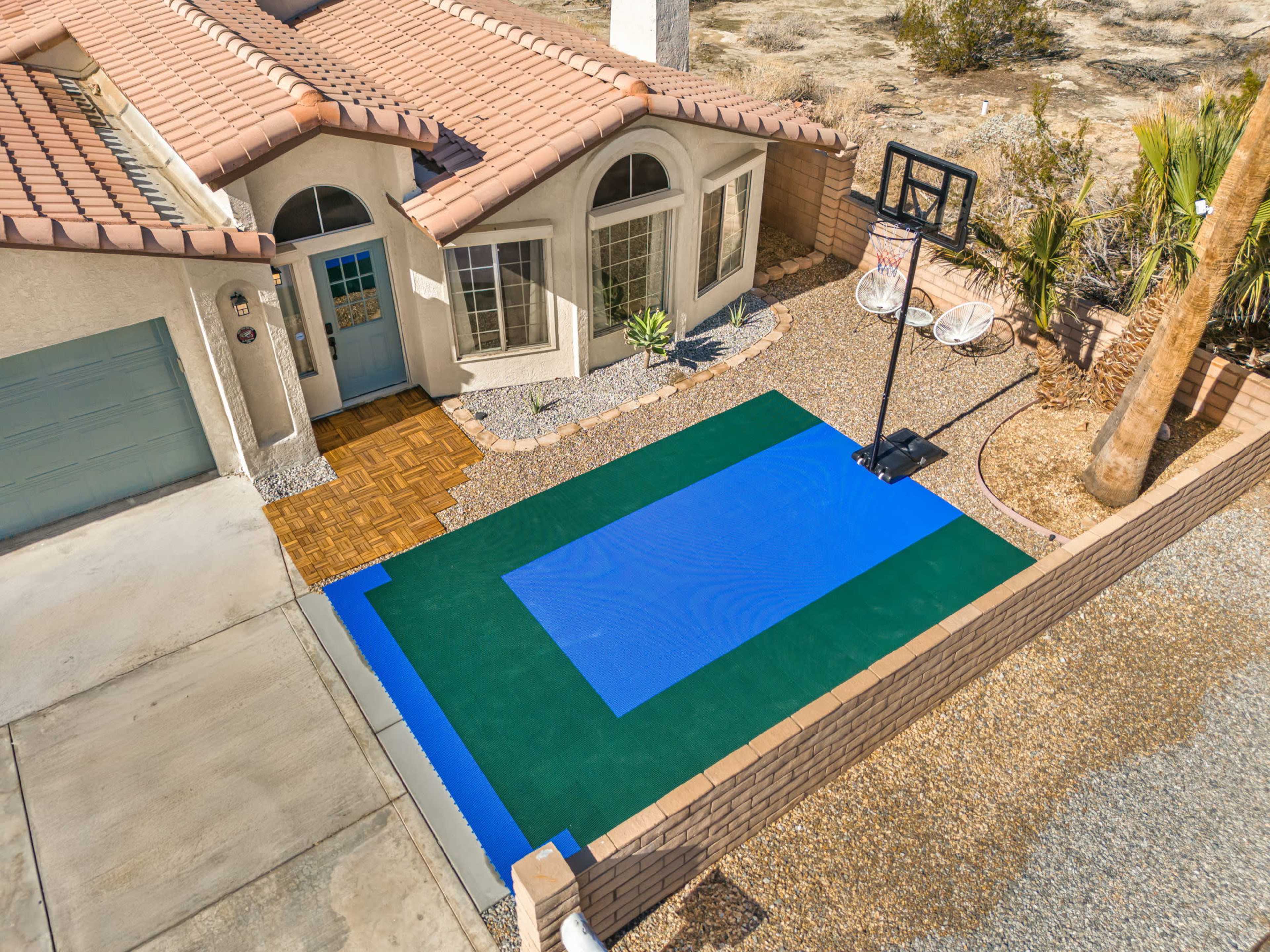 A residential driveway features a blue and green basketball court with a hoop adjacent to a house with a tiled roof.