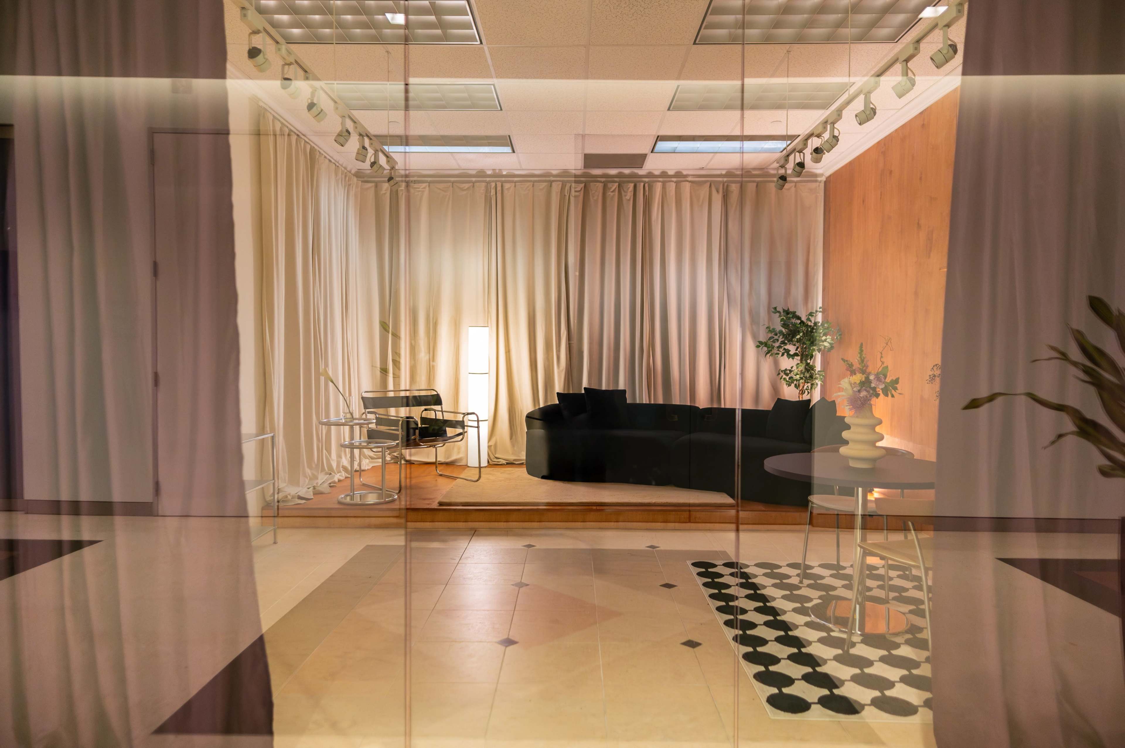 The image shows a modern interior space with a black sofa, a round table, a patterned rug, and curtains dividing the area.