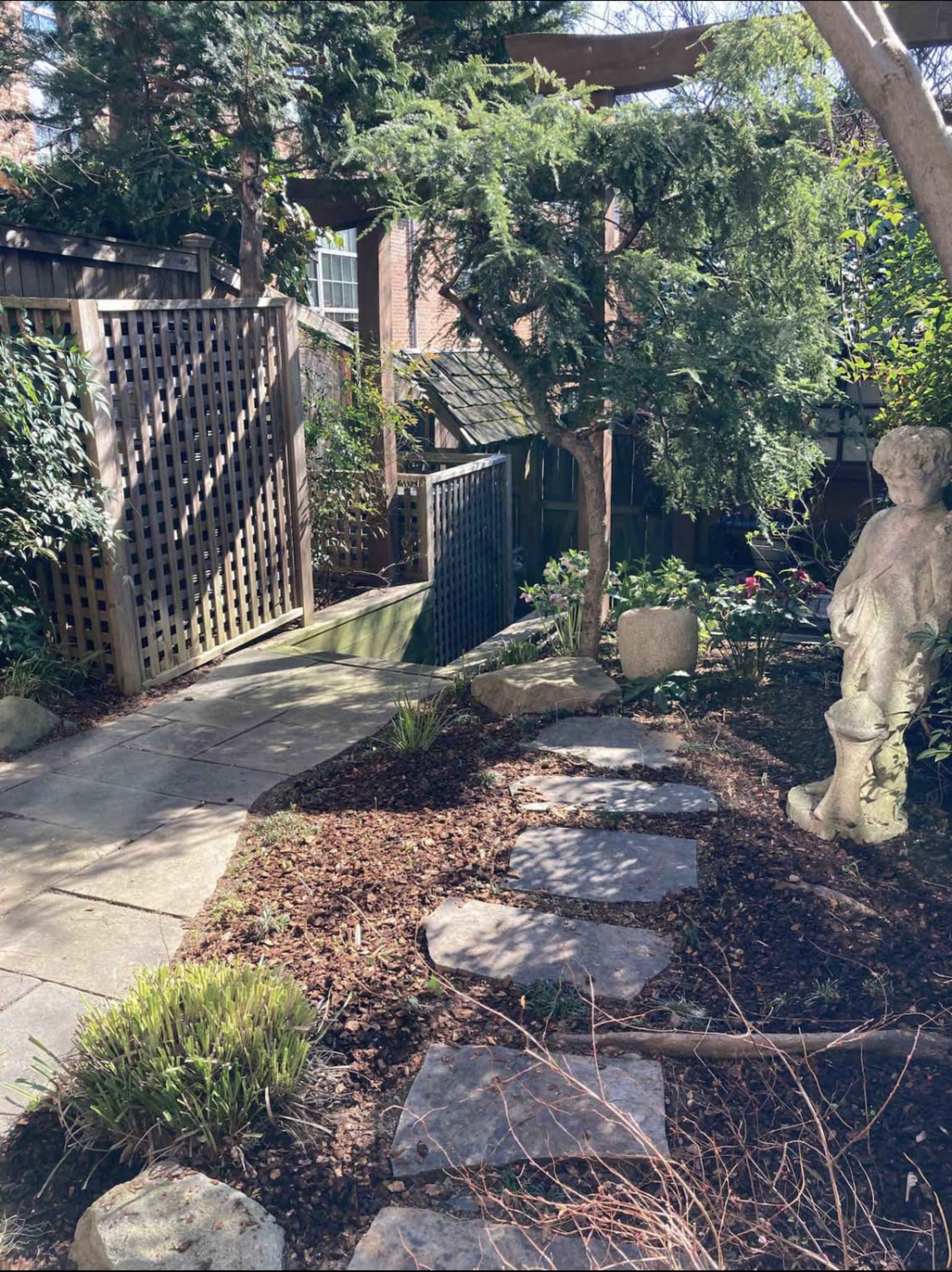 A stone path winds through a garden featuring a sculpture, greenery, and wooden fences on either side.