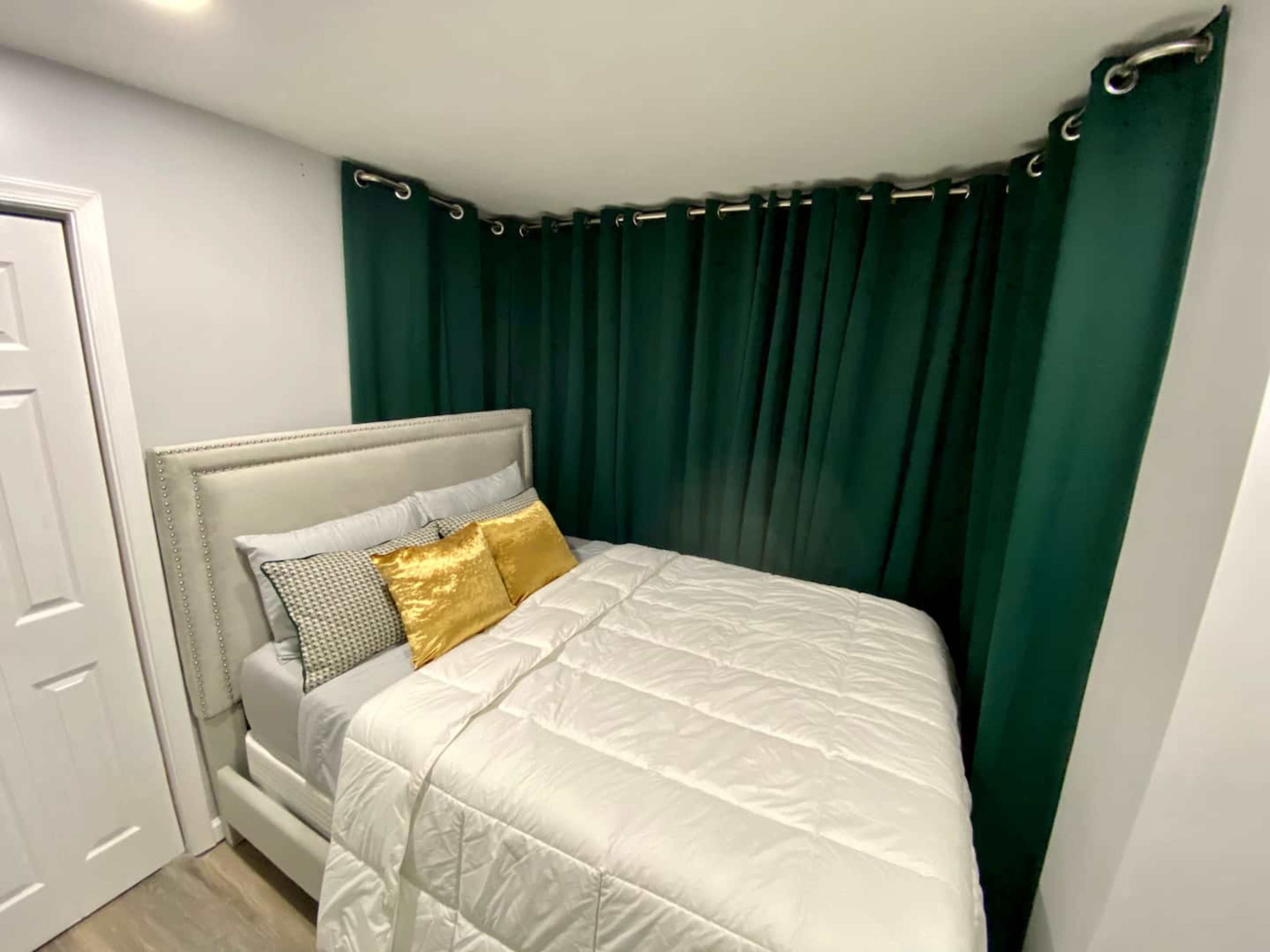 A small bedroom with a bed covered in a white quilt and adorned with patterned and gold pillows, surrounded by dark green curtains.
