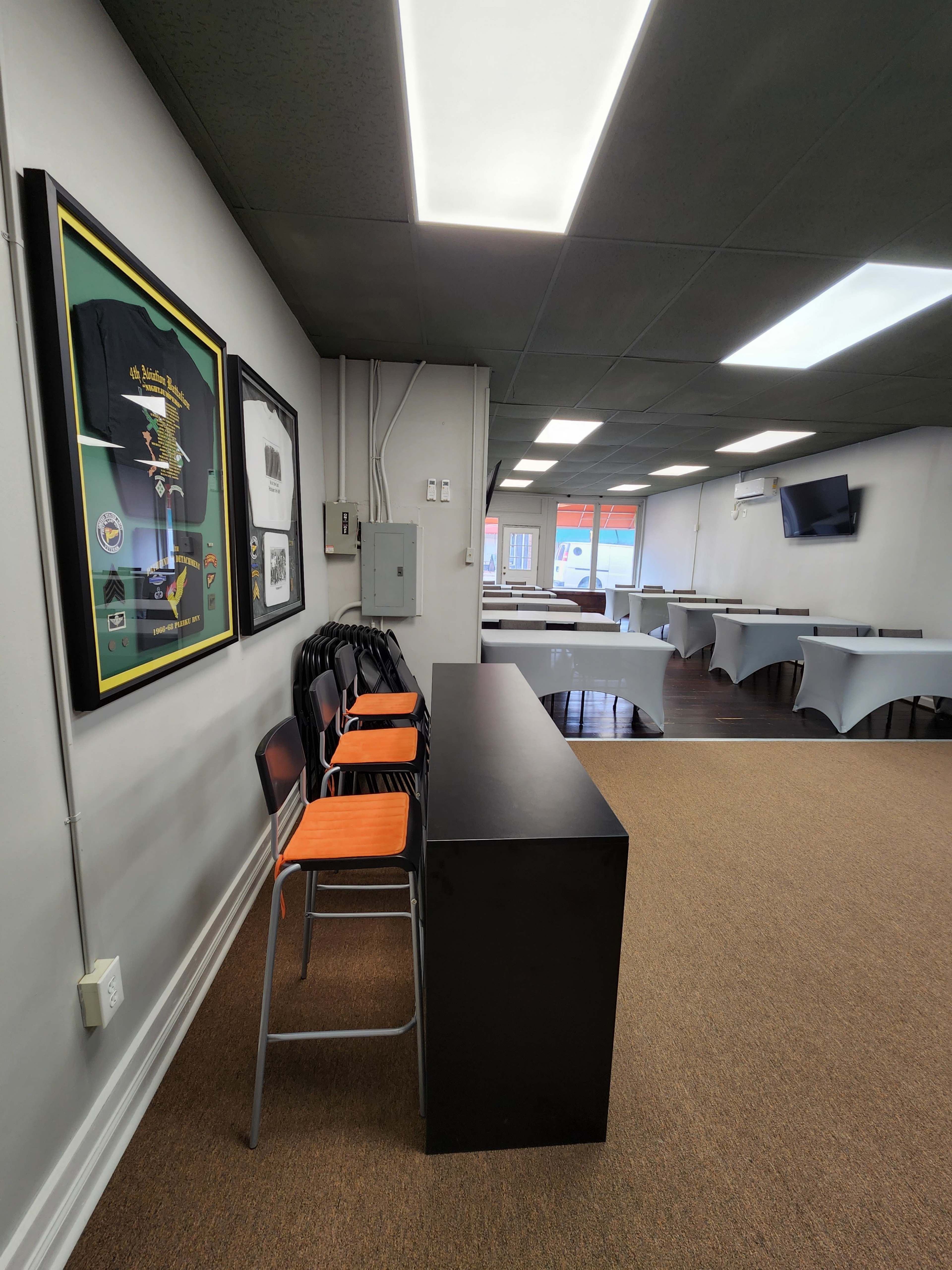 An empty room with several tables covered in gray cloth, a dark countertop with orange chairs stacked on one side, and framed artwork on the wall.