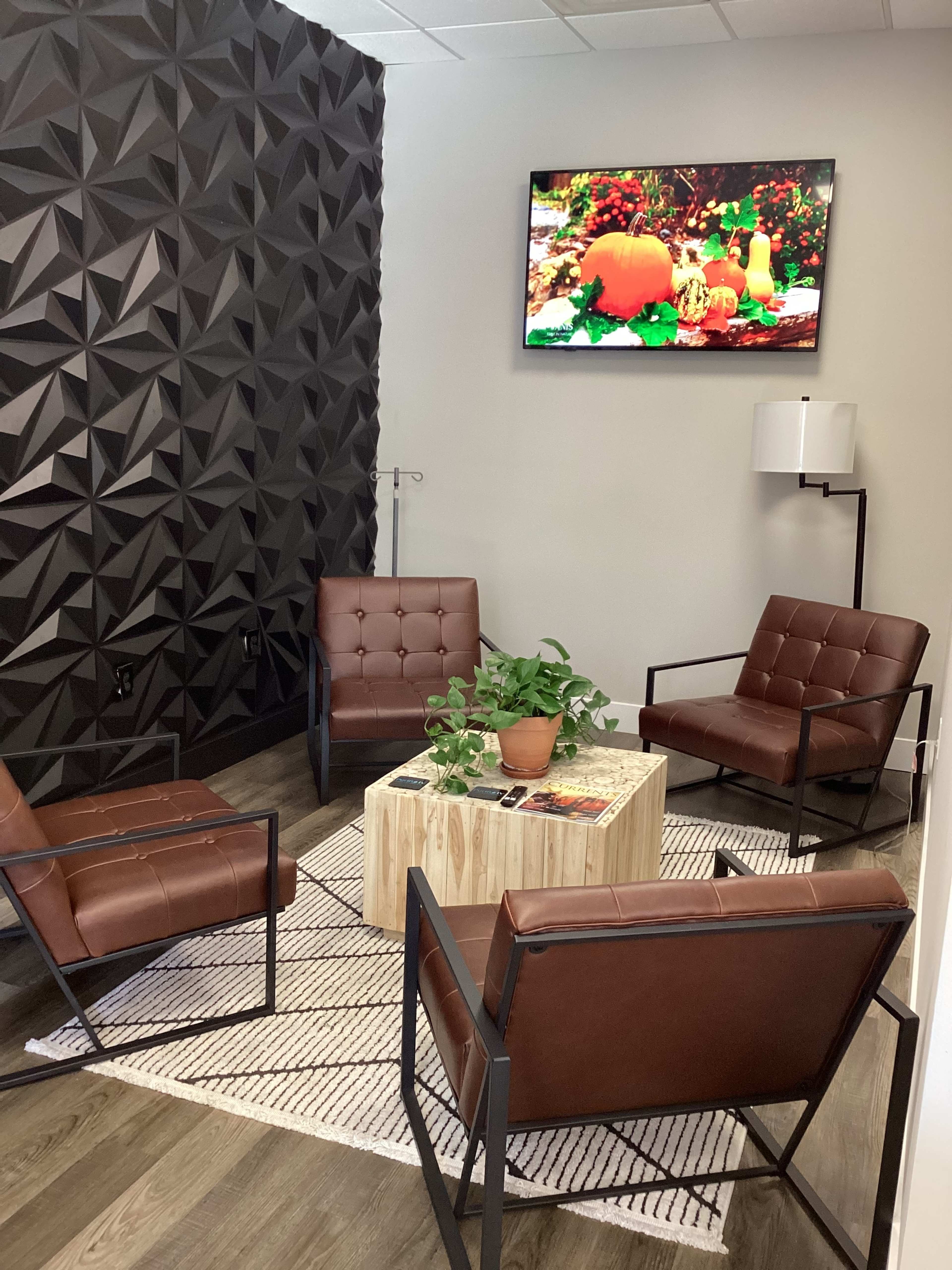 A modern waiting area with four brown leather chairs arranged around a wooden table, featuring a plant and a wall-mounted television displaying a colorful scene.