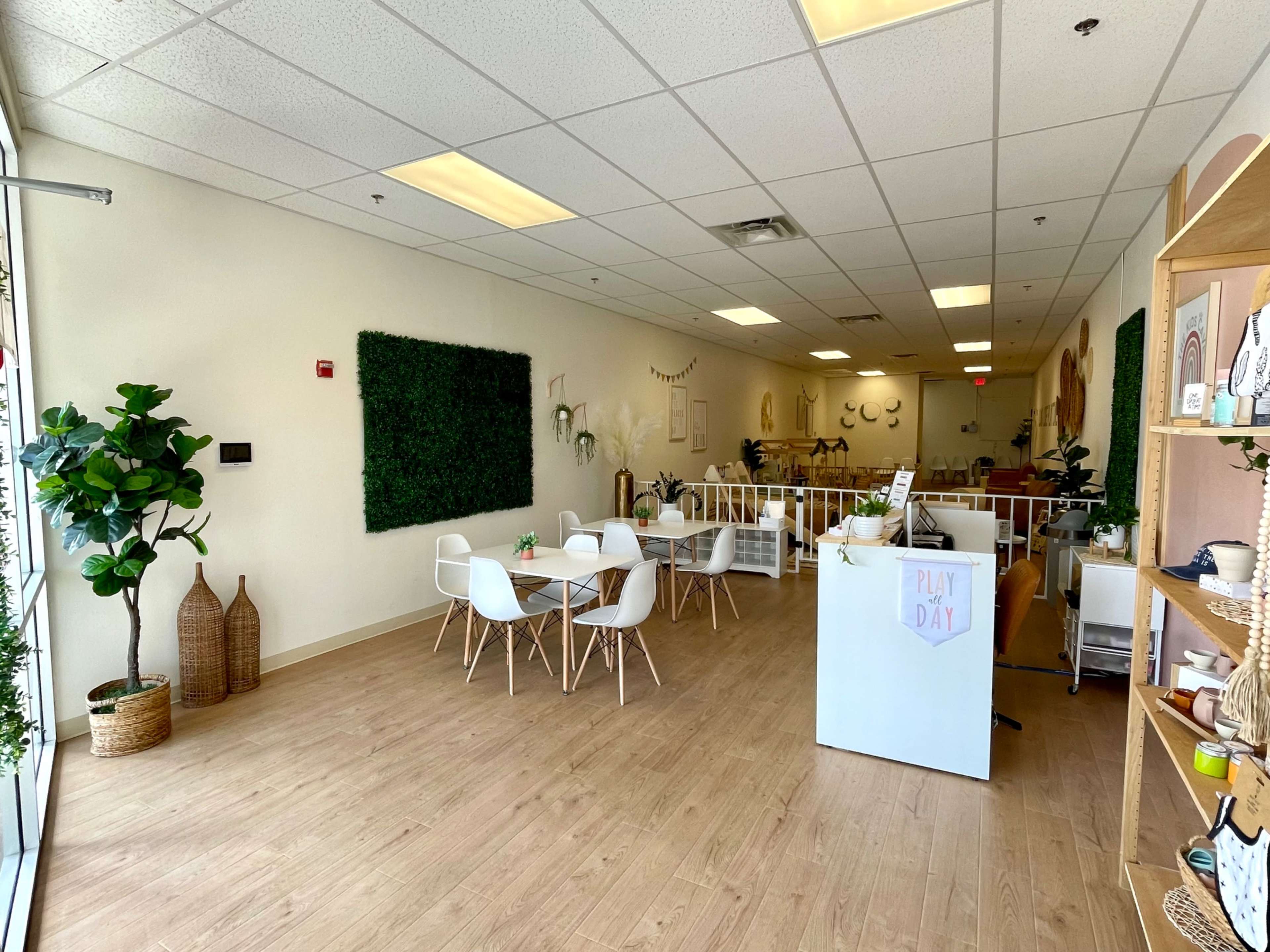 The image shows a spacious, bright interior of a room with wooden flooring, featuring a seating area with white chairs and tables, green wall decorations, and shelves displaying various items.
