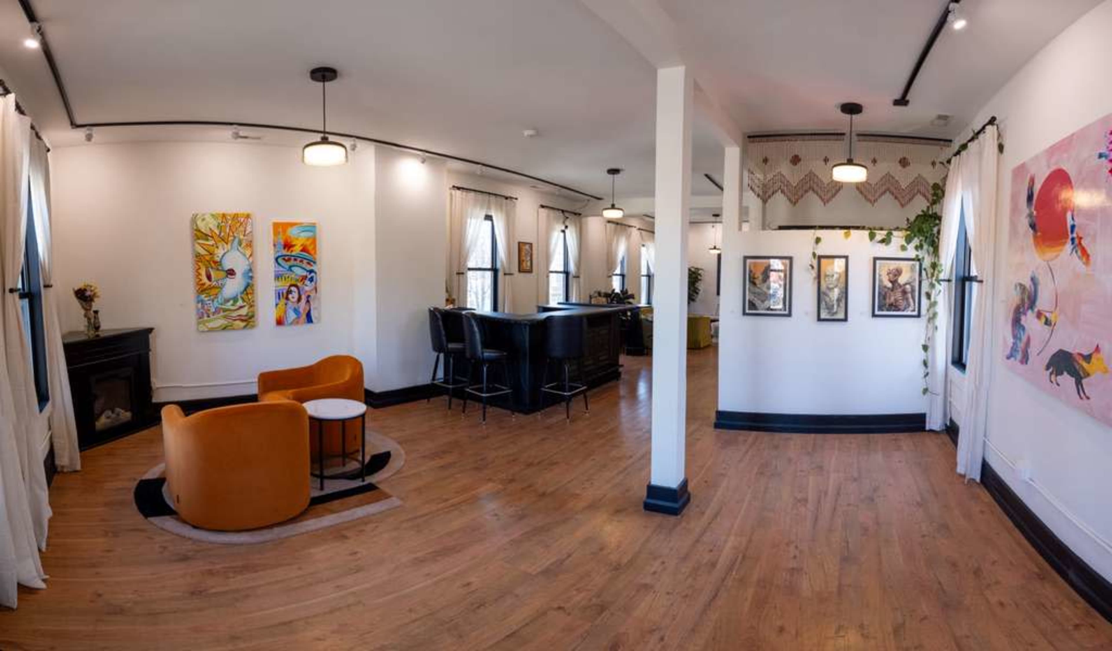 The image shows a spacious, well-lit interior room with two orange chairs and a small table, framed artwork on the walls, and a dark bar area in the background.