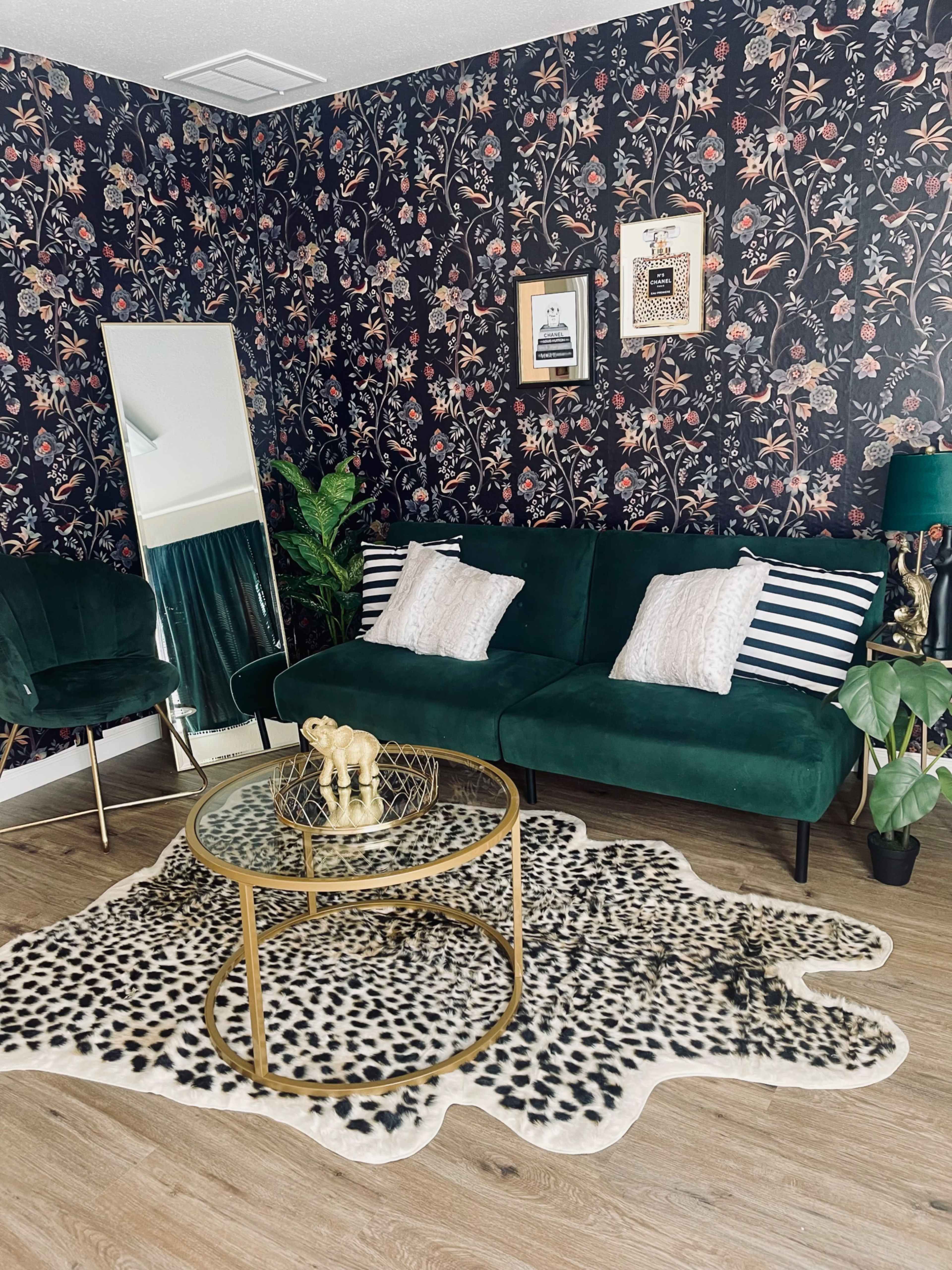 The image shows a stylish room with floral wallpaper, a green velvet sofa, a round coffee table, a large mirror, and a decorative rug.