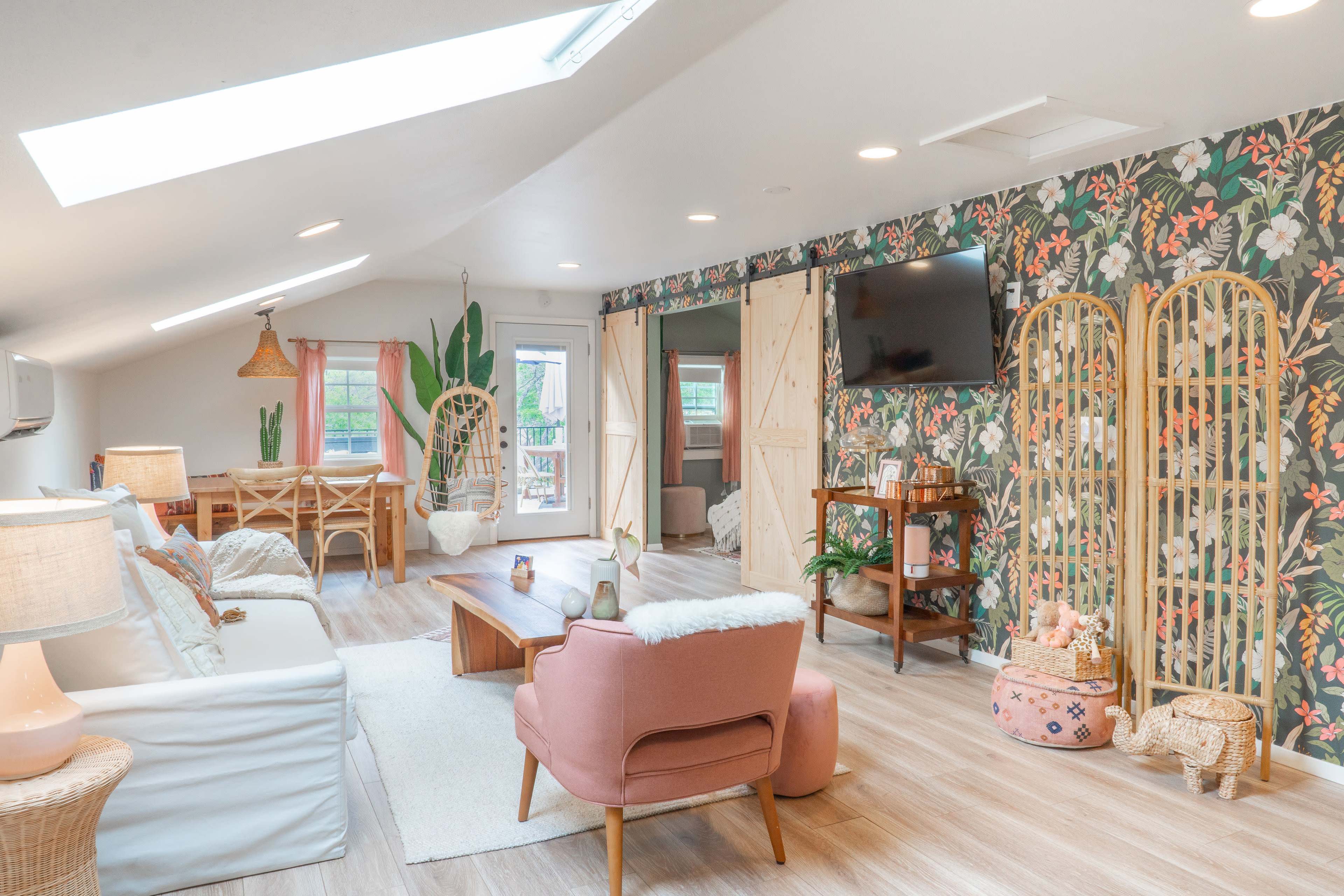 A bright, modern living room with floral wallpaper, a light-colored sofa, a pink armchair, and wooden furniture, featuring large windows and skylights.