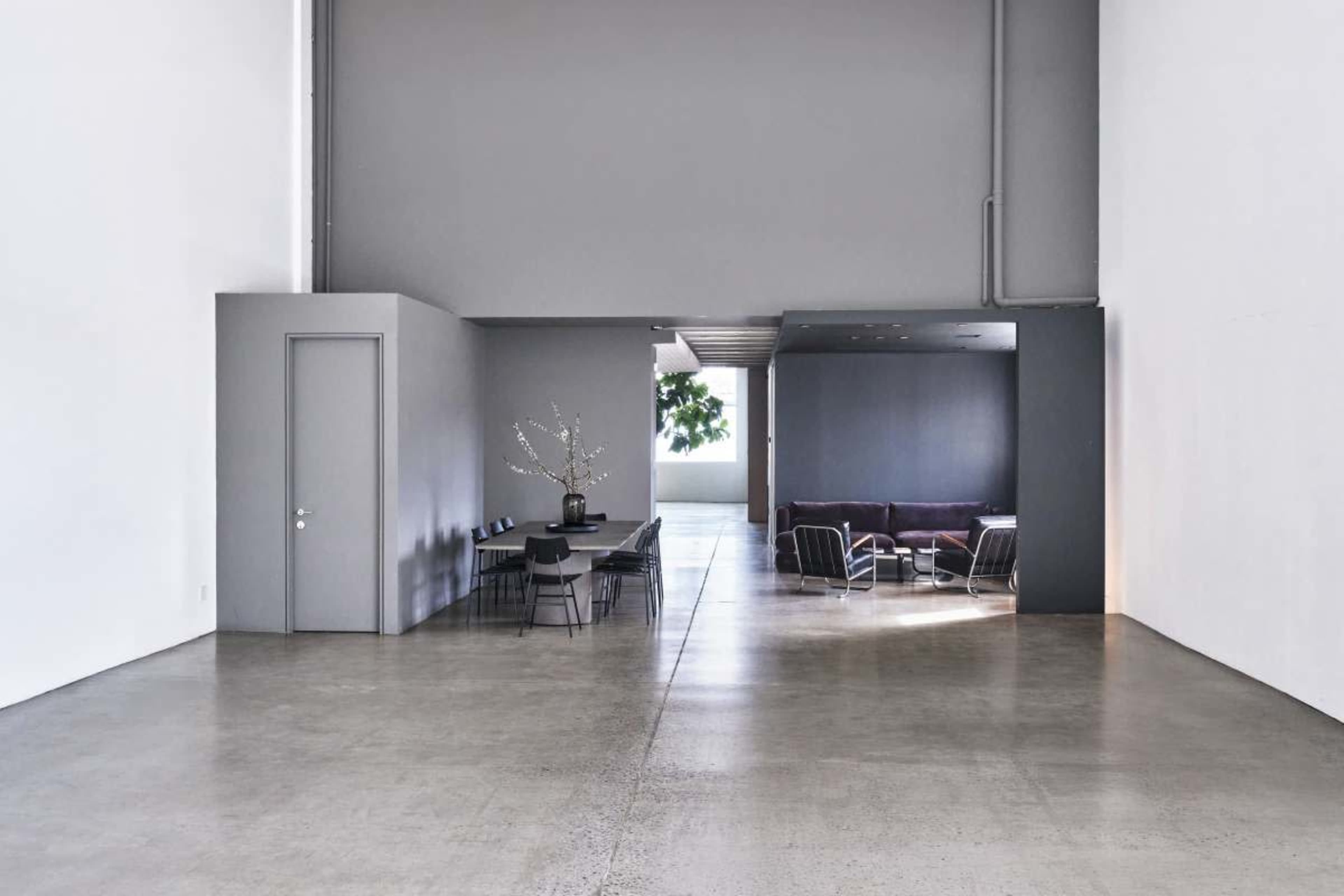 The image displays a spacious, modern interior with a grey color palette, featuring a dining area with a table and chairs on one side and a seating area with a sofa on the other.