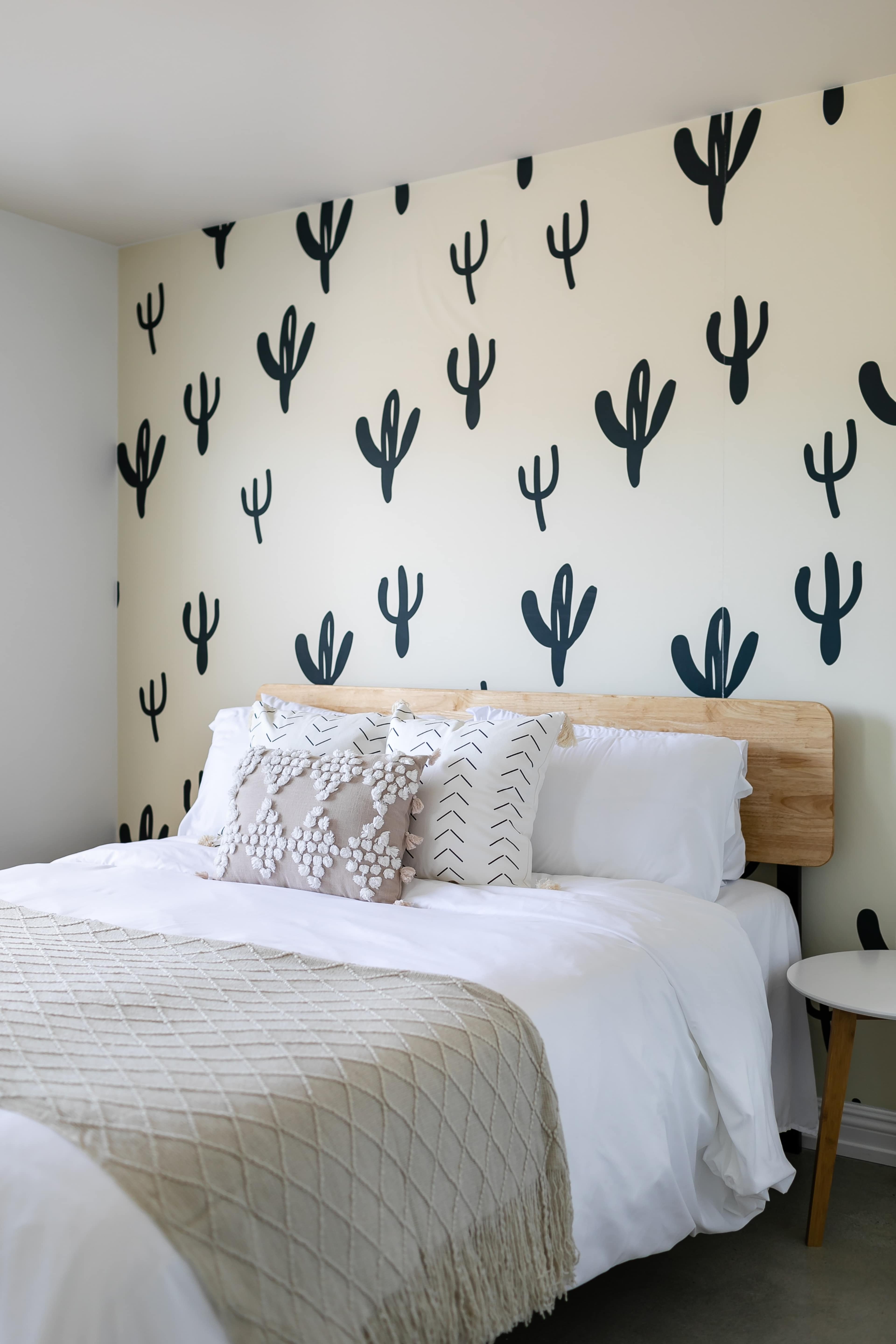 A neatly arranged bedroom with a bed featuring white linens and a wooden headboard against a wall decorated with black cactus patterns.