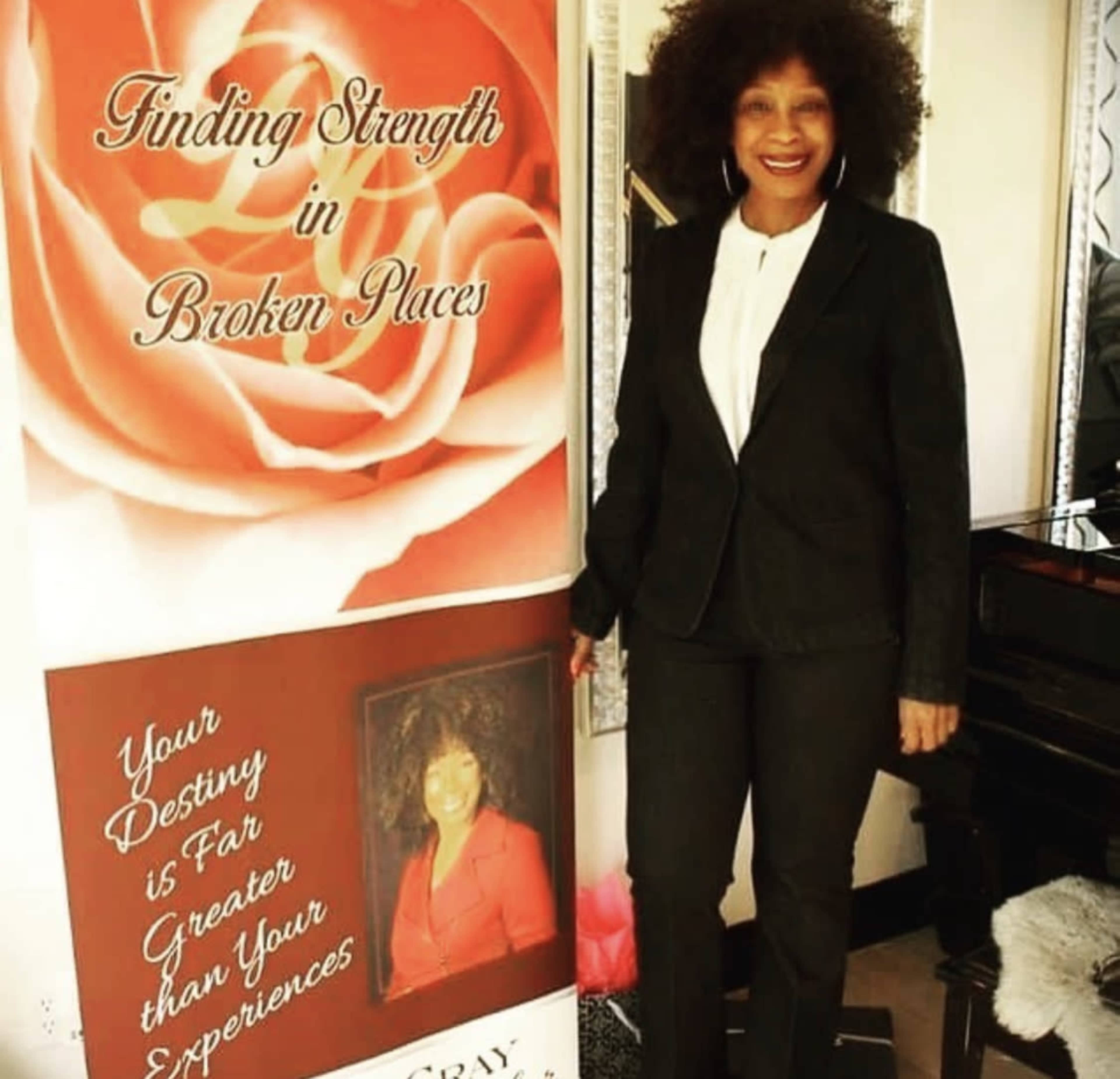 A woman stands beside a banner that reads "Finding Strength in Broken Places," in a room with a piano and decorated with a rose graphic.