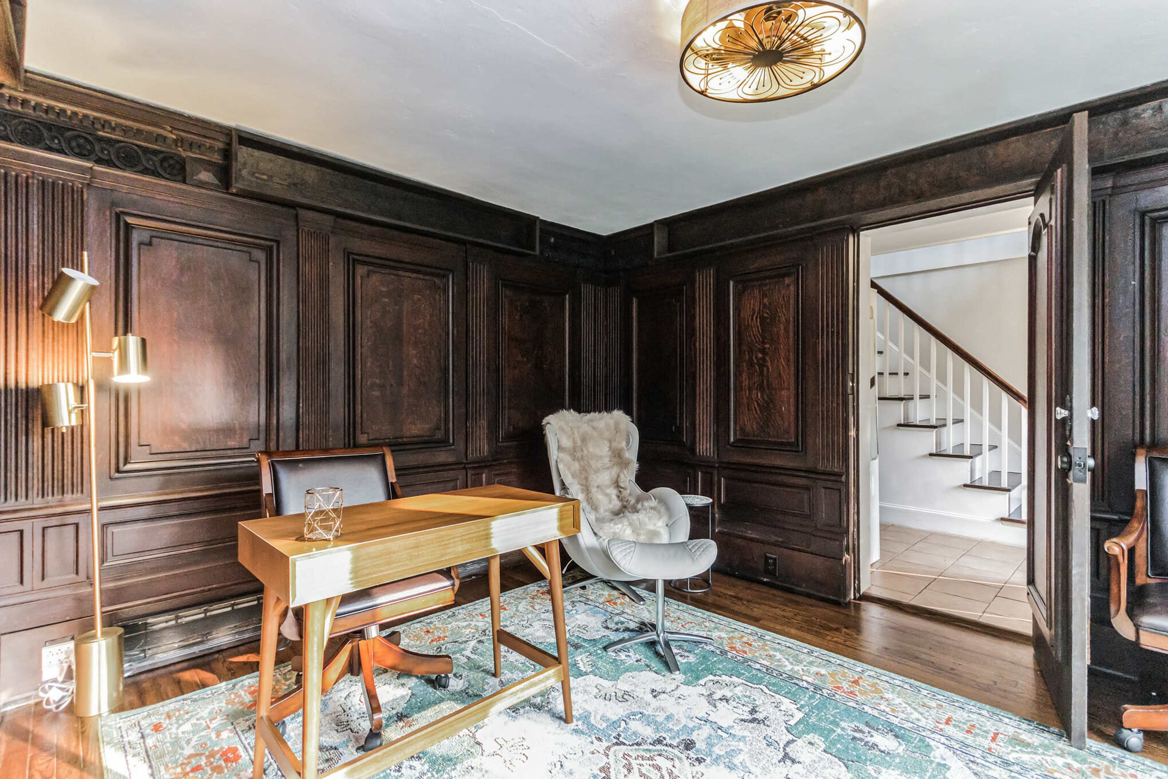 The image depicts a home office with dark wooden paneled walls, a wooden desk, a white chair, a light rug, and a staircase visible through an open door.