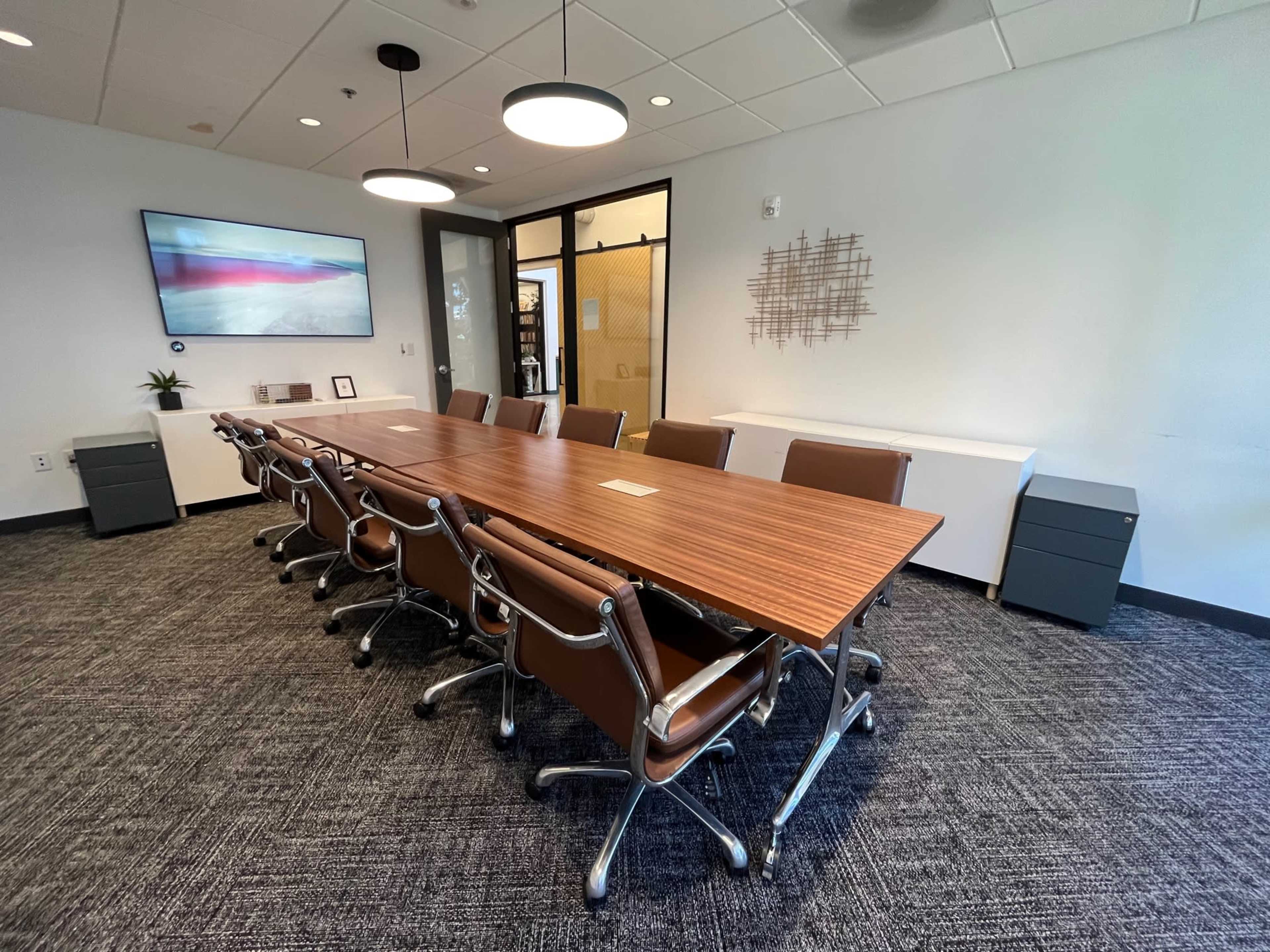 A large conference room features a long wooden table with eight rolling chairs, a television on the wall, and two filing cabinets in the corner.
