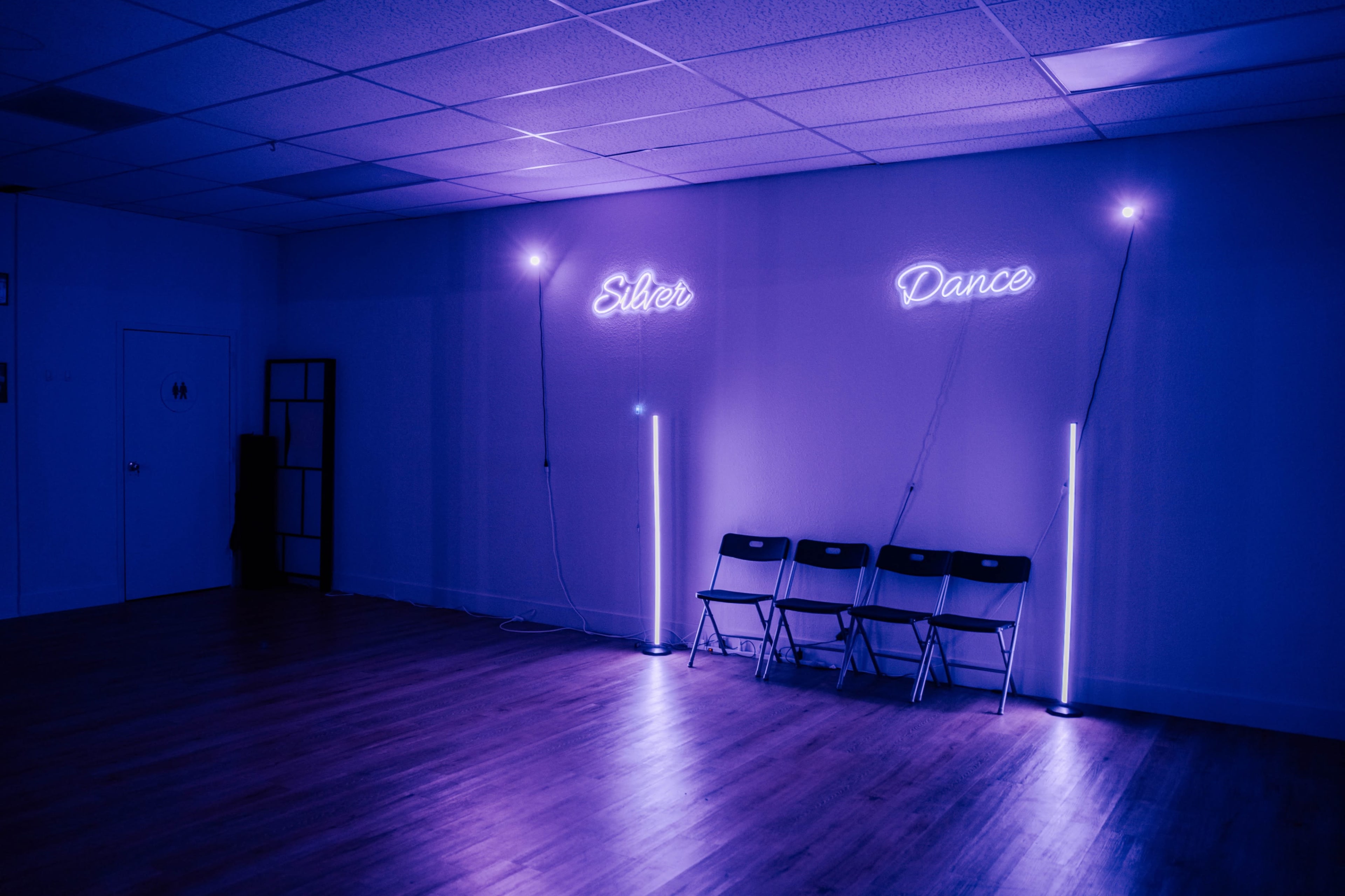 The image shows an empty dance studio with purple neon signs that read "Silver" and "Dance" along the wall and several black chairs aligned in front.