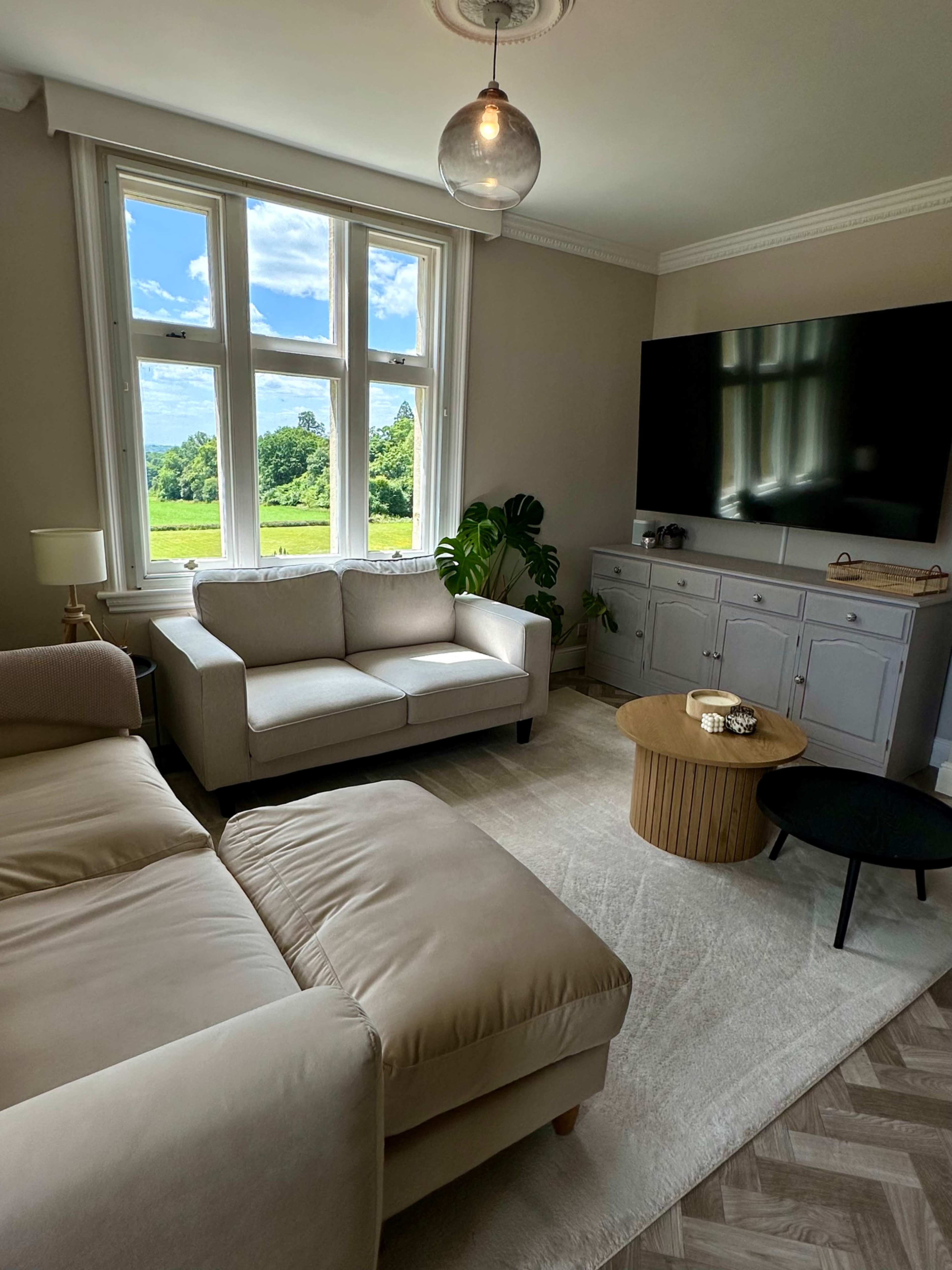 A bright living room features a light-colored sofa, a wooden coffee table, and a large television mounted on the wall, with large windows overlooking a green landscape.