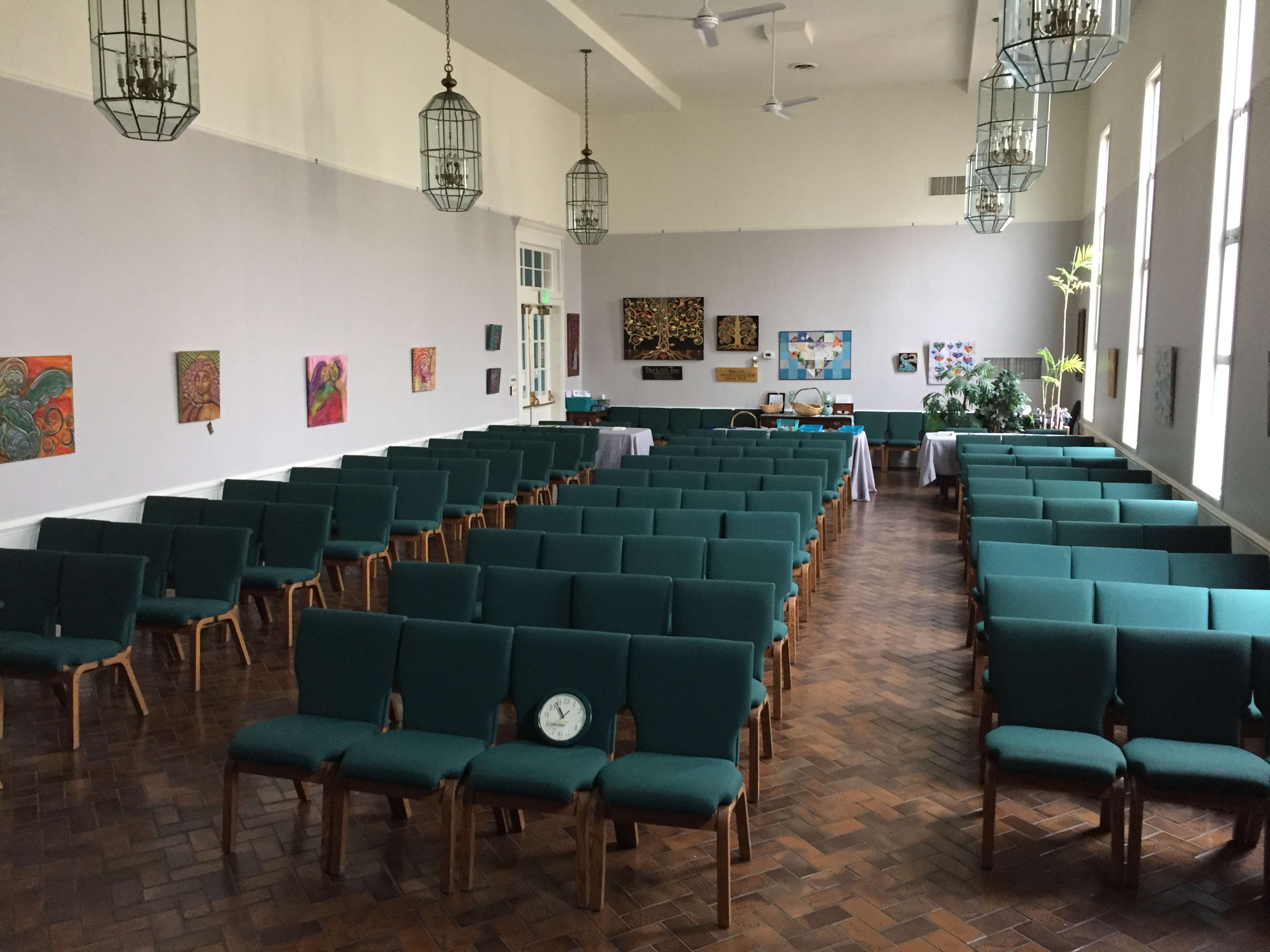 A large, empty room arranged with rows of green chairs facing a stage, decorated with paintings on the walls and featuring windows that allow natural light.