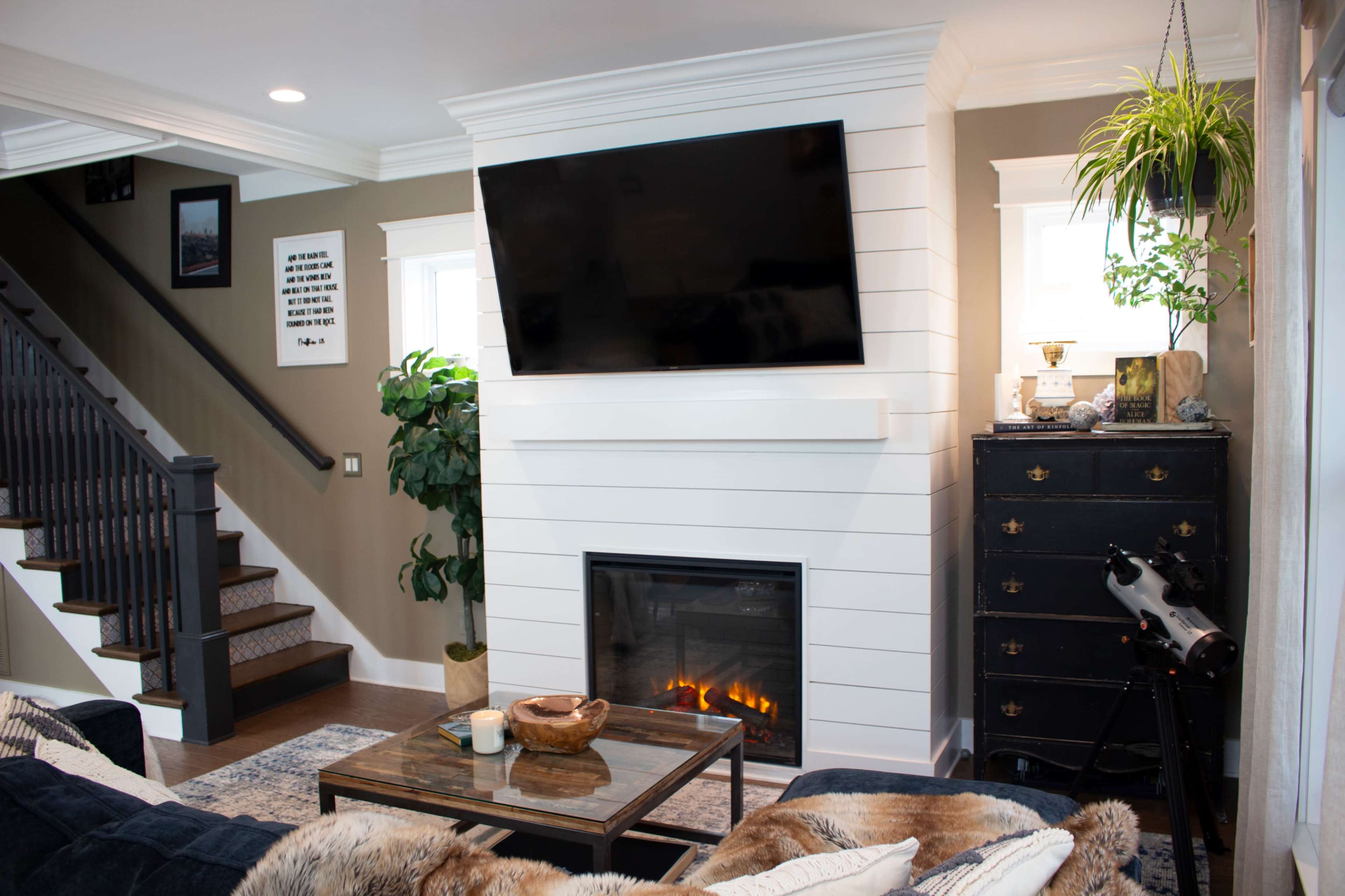The image shows a cozy living room with a mounted flat-screen TV above a white shiplap fireplace, a dark dresser, a green potted plant, and a staircase in the background.
