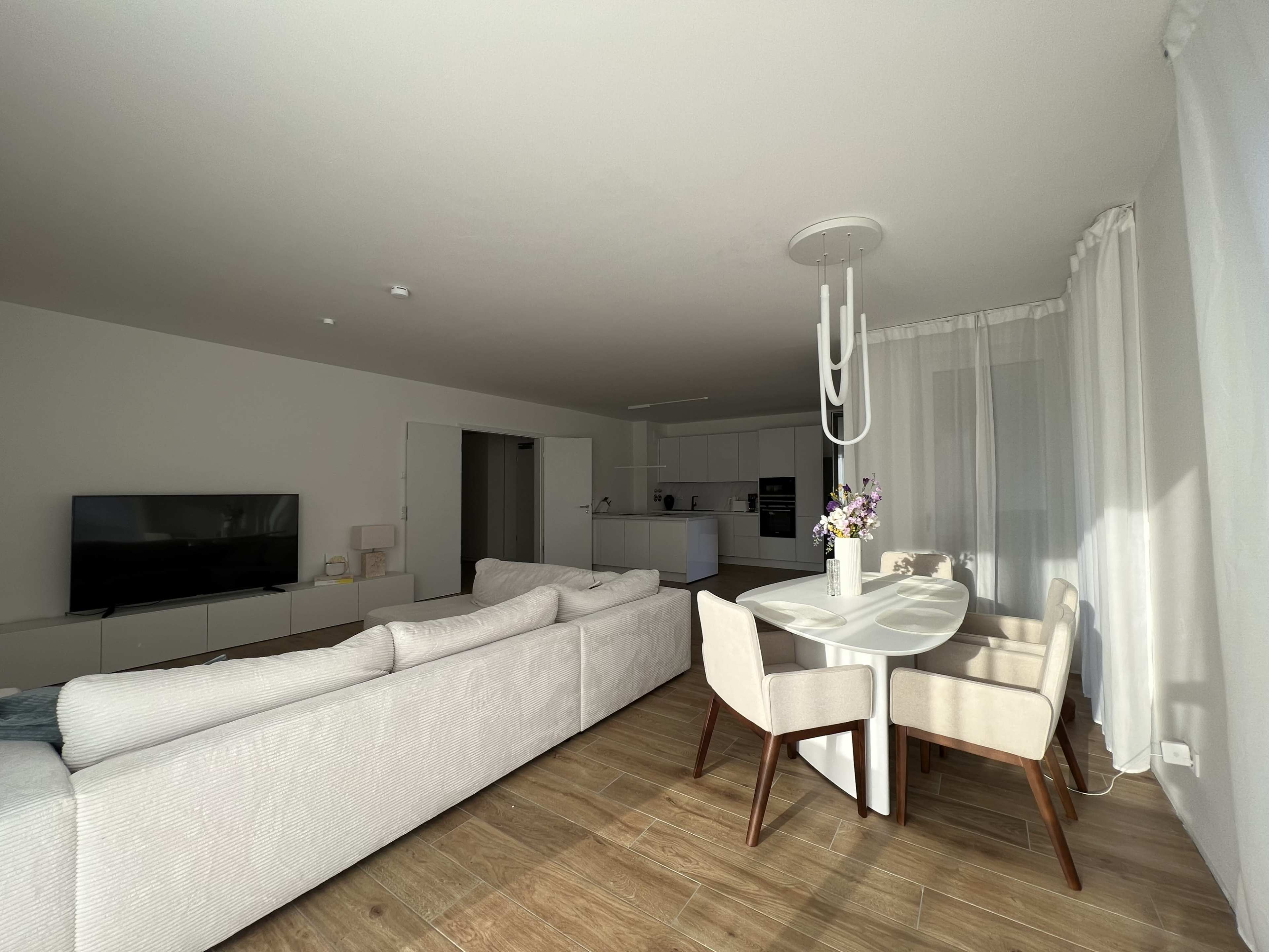 The image shows a modern living space featuring a light-colored sofa, a dining table with chairs, and a kitchen area in the background.