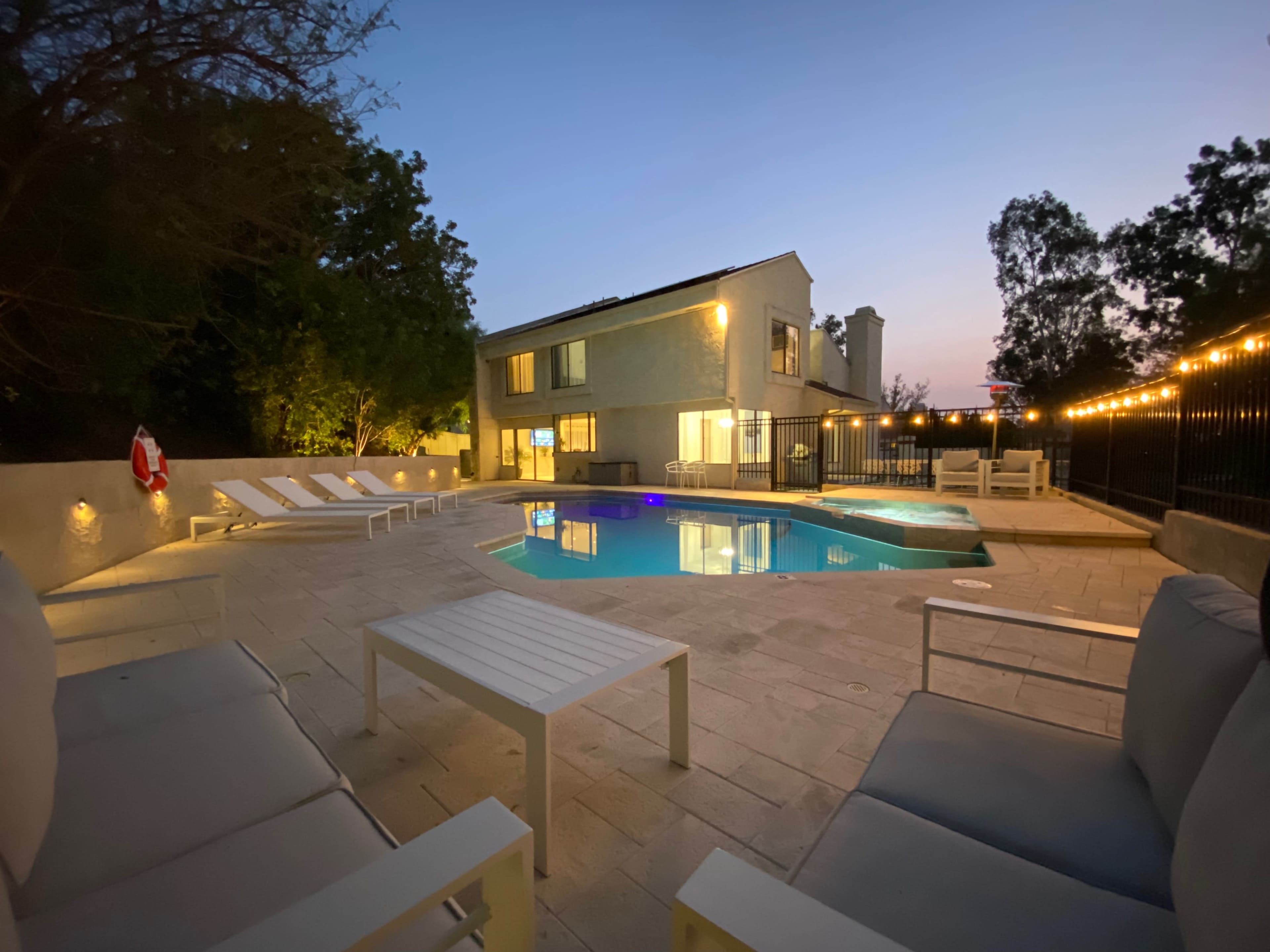 A modern two-story house overlooks a swimming pool surrounded by lounge chairs and illuminated by string lights during twilight.