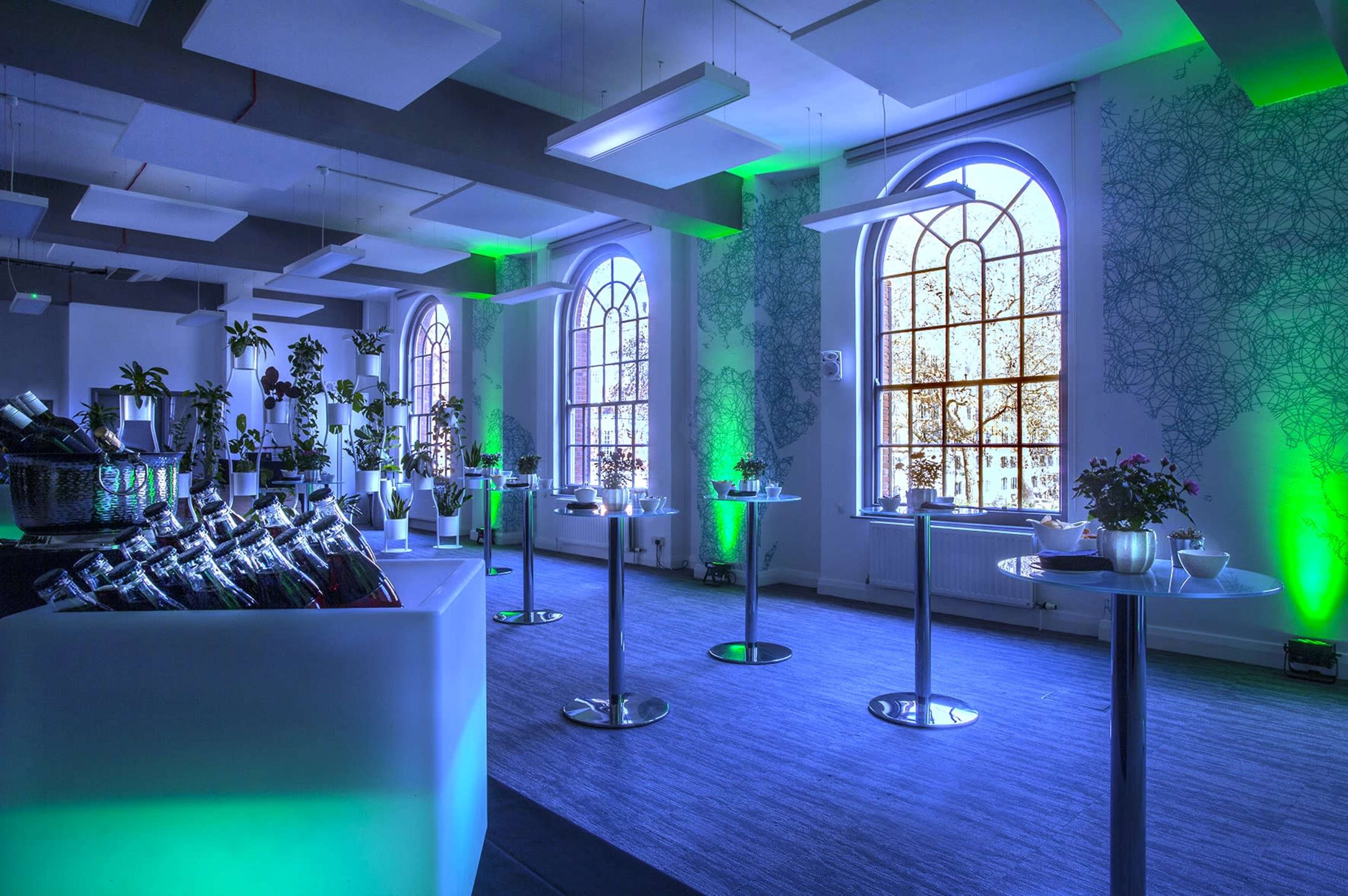 A well-lit event room features tables with food arrangements and plants, illuminated by green lighting and large windows.