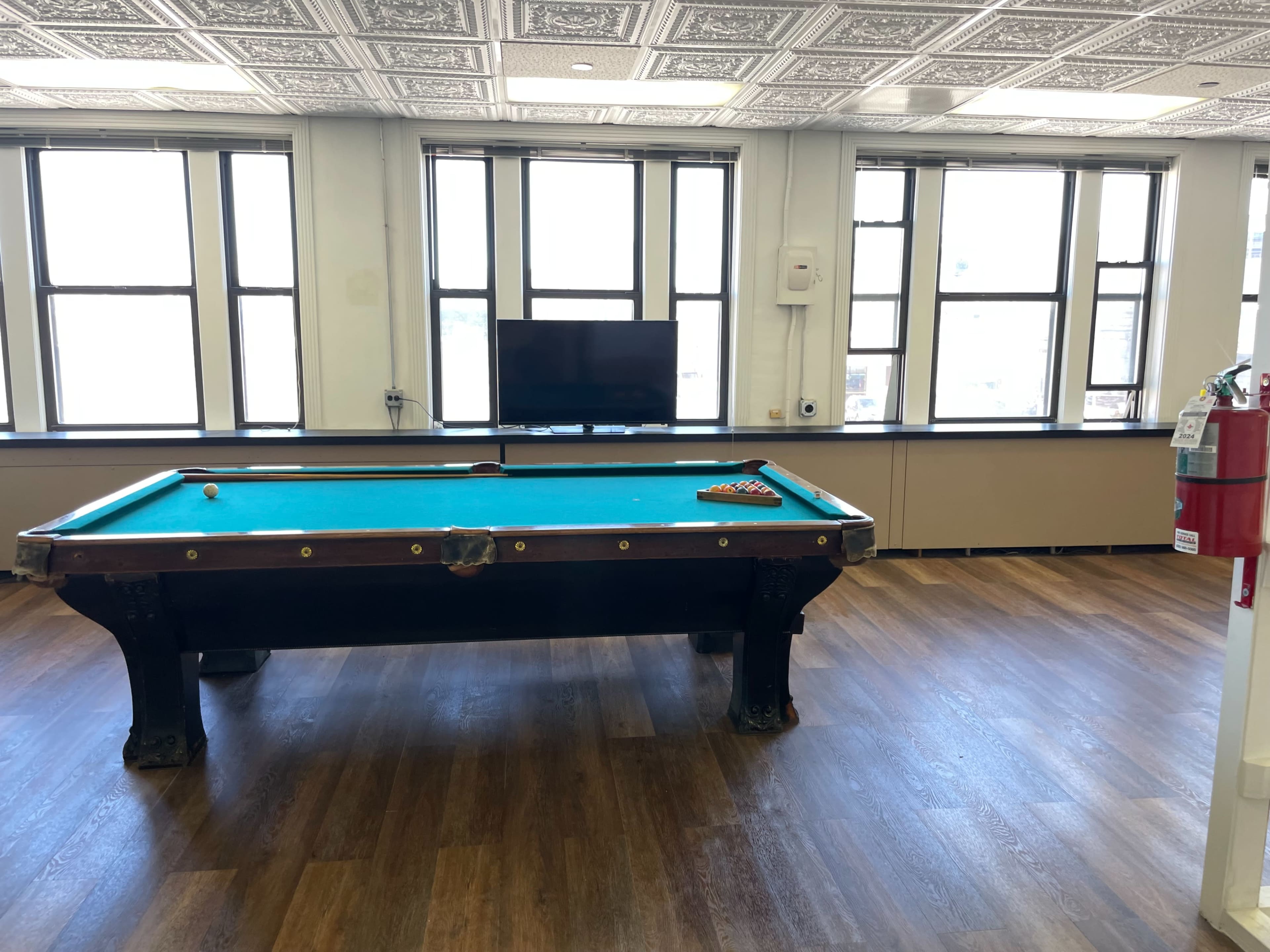 The image shows a pool table with a blue felt surface, located in a well-lit room featuring large windows and wooden flooring.