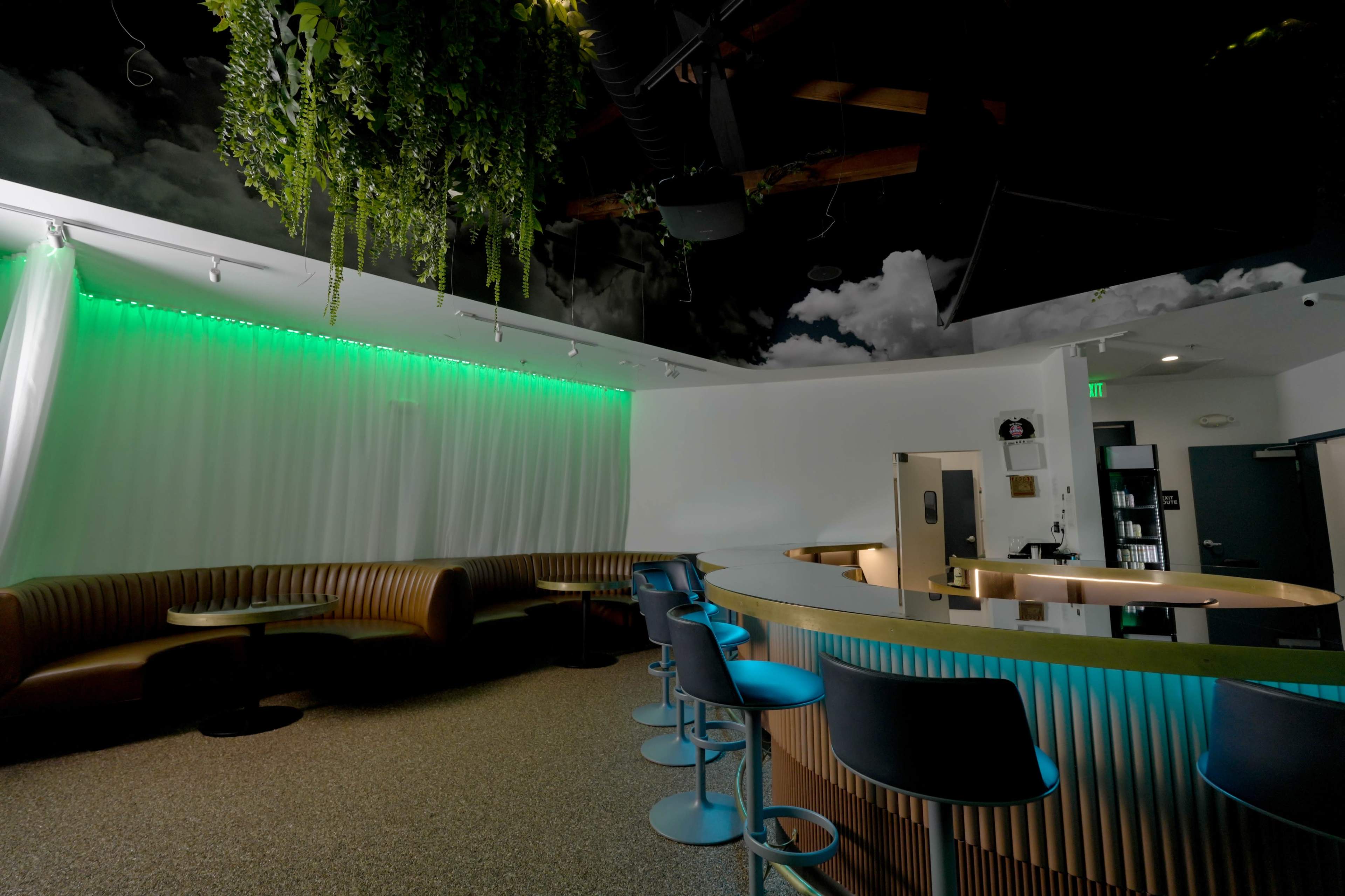 The image shows a modern lounge area featuring a curved bar with high stools, a seating area with a leather couch, and ambient green lighting.