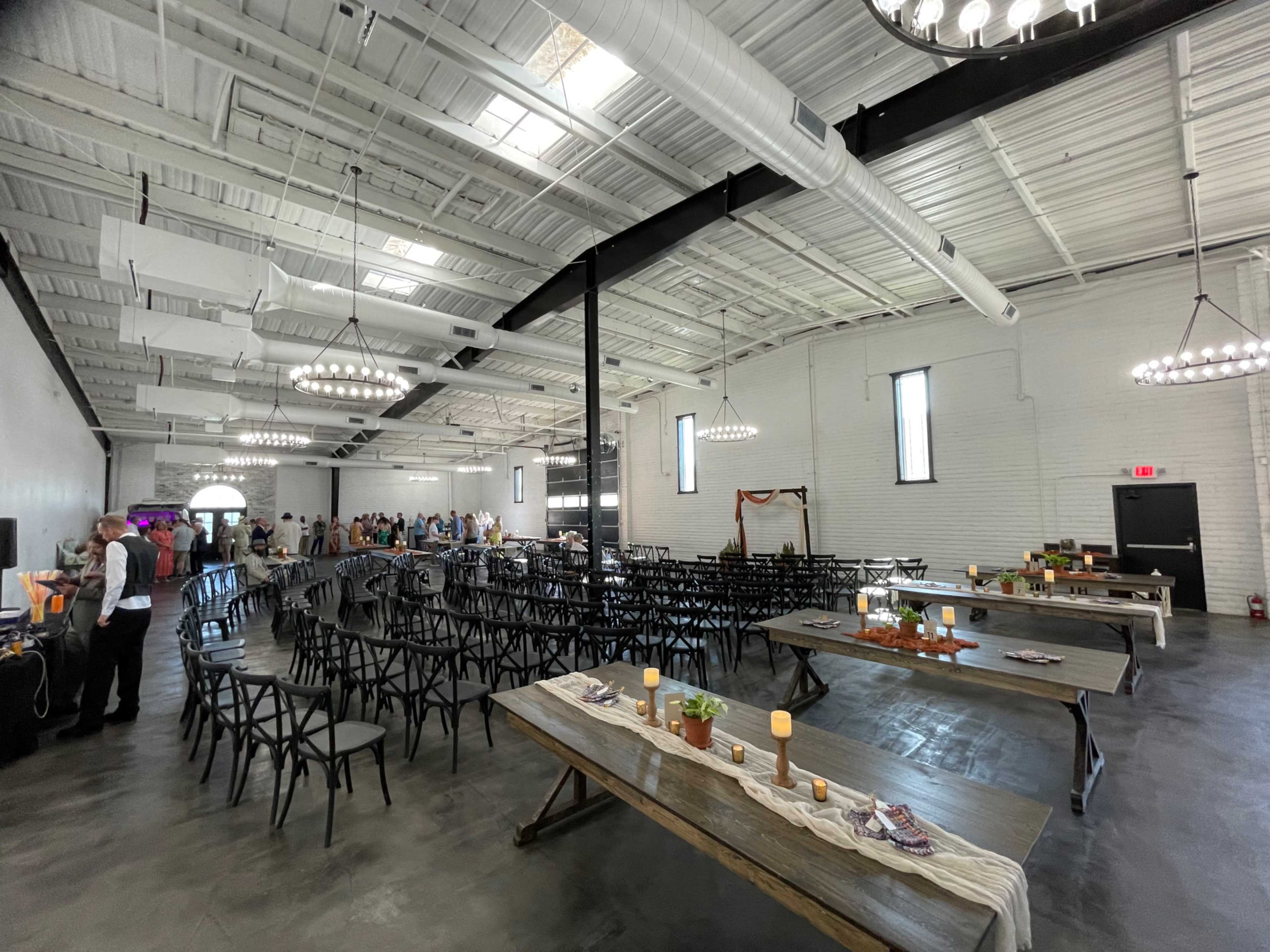 The image shows a large, industrial-style event space set up with rows of black chairs and long wooden tables, with guests mingling in the background.