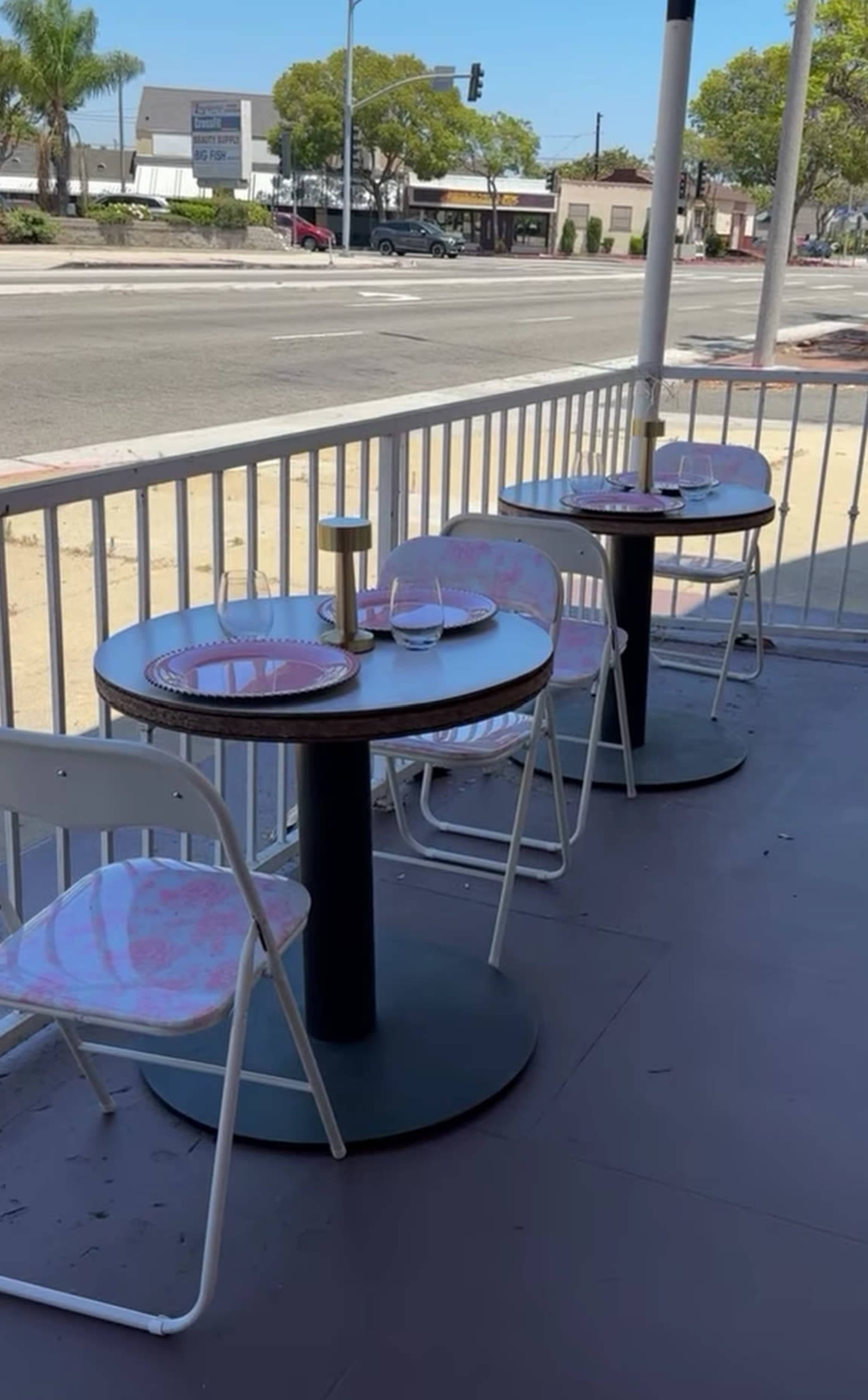 The image shows a shaded outdoor dining area with two round tables set for meals, each featuring plates and glasses, alongside a busy street in the background.