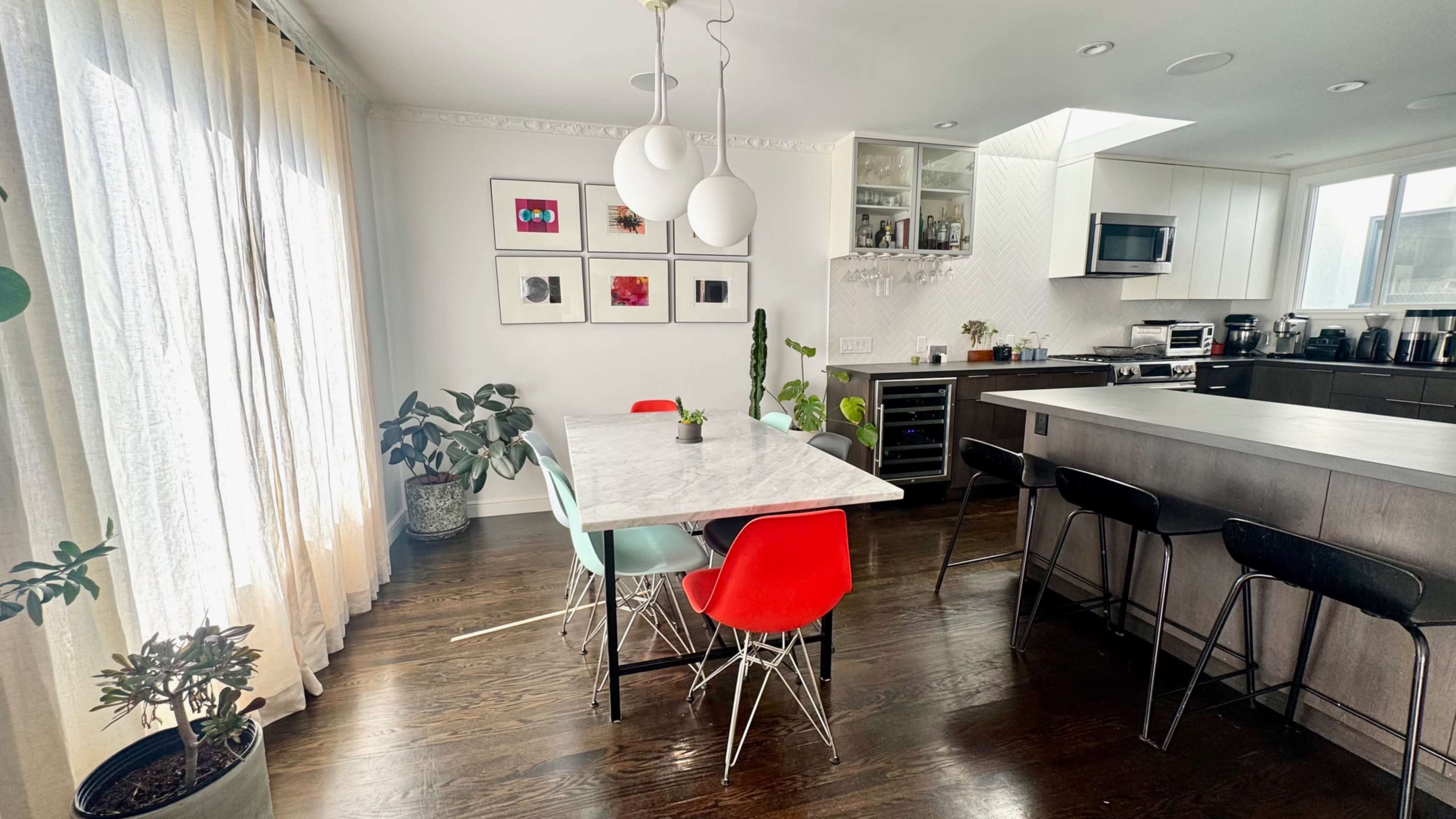 A modern kitchen-dining area features a marble table surrounded by colorful chairs, large windows with sheer curtains, and sleek cabinetry.
