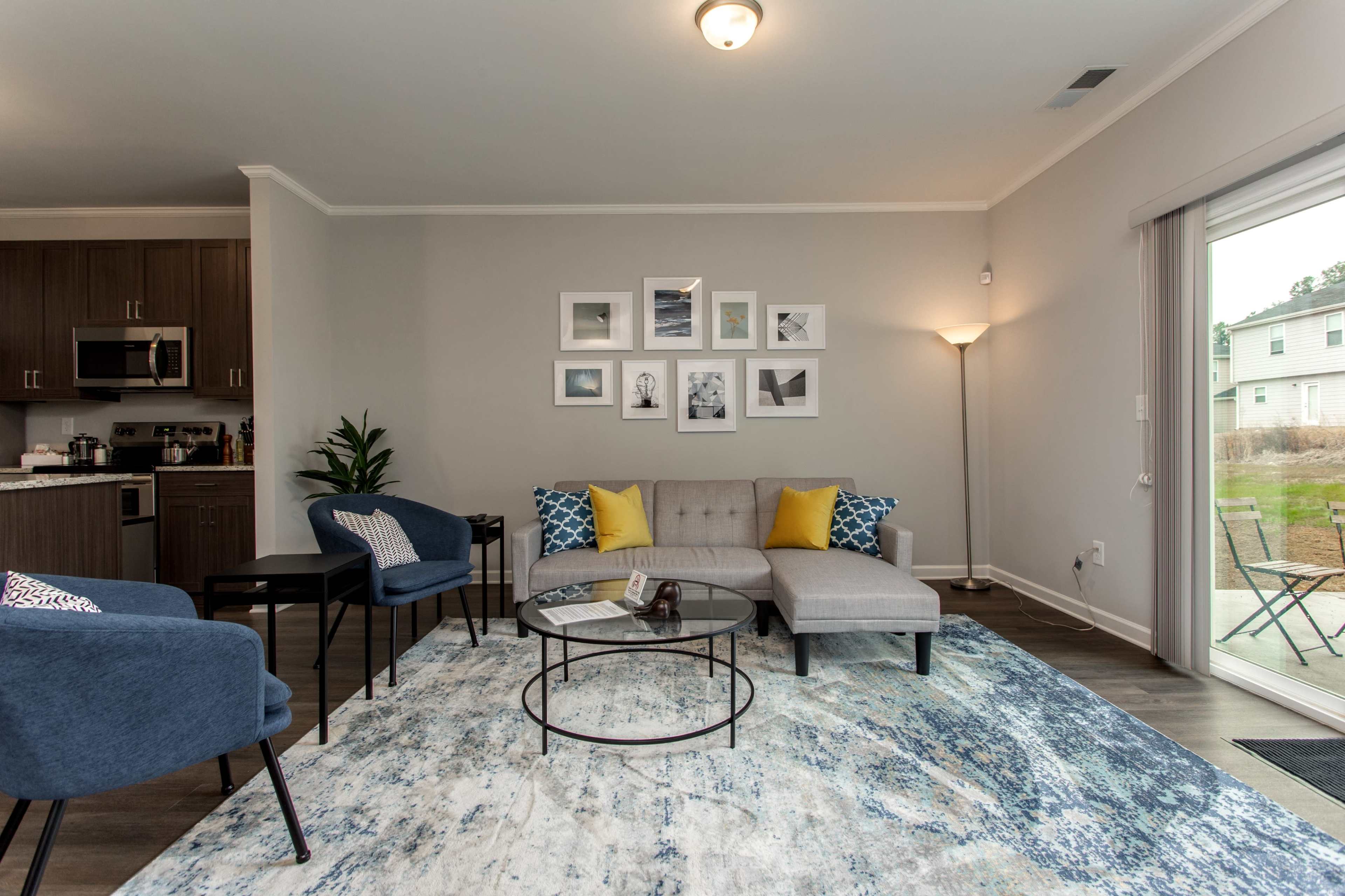 A modern living room features a gray sofa, decorative armchairs, framed artwork on the wall, and a glass coffee table atop a patterned rug.