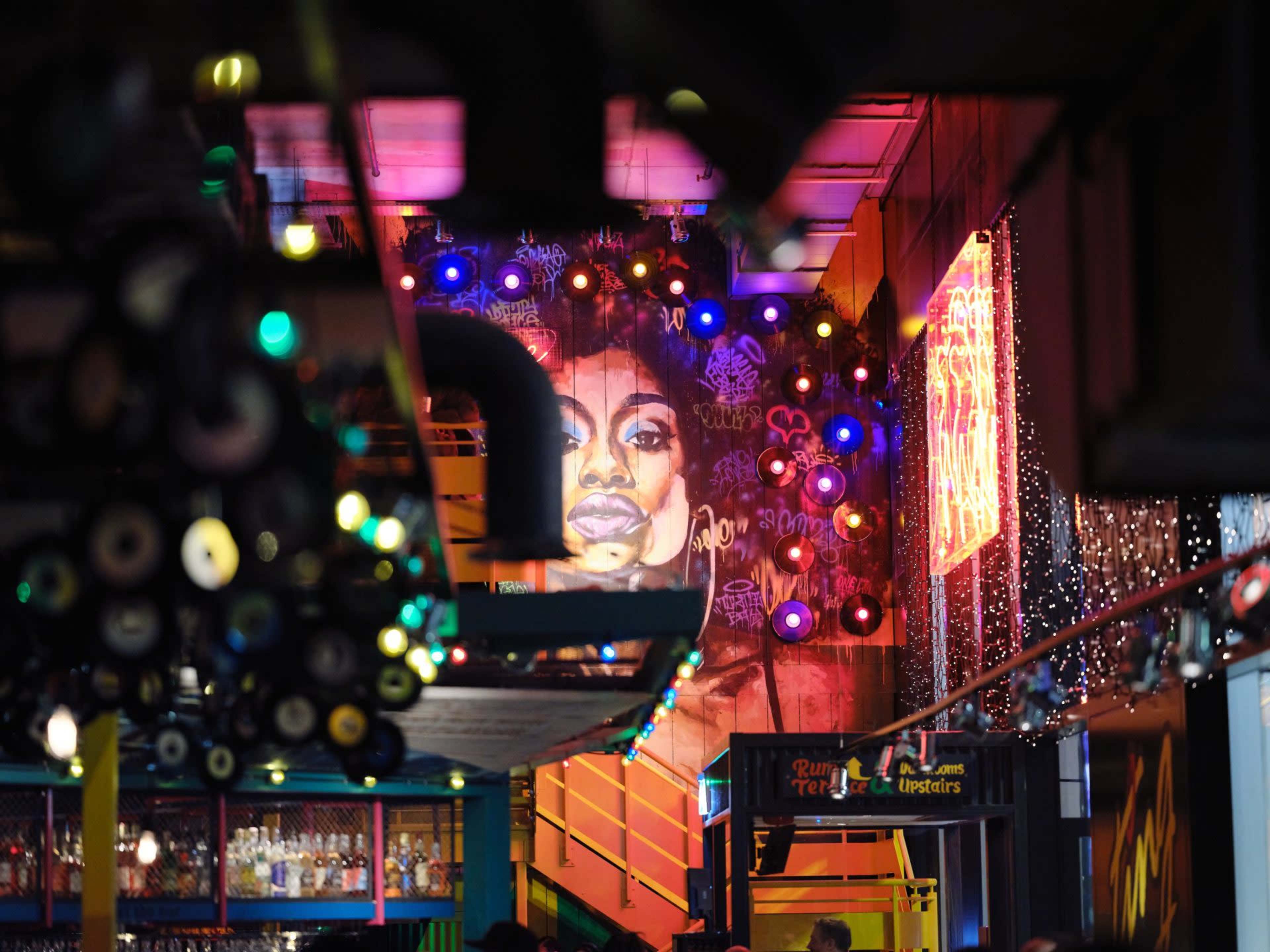 The image shows an indoor space with vibrant murals and colorful lights, featuring a large portrait on the wall.