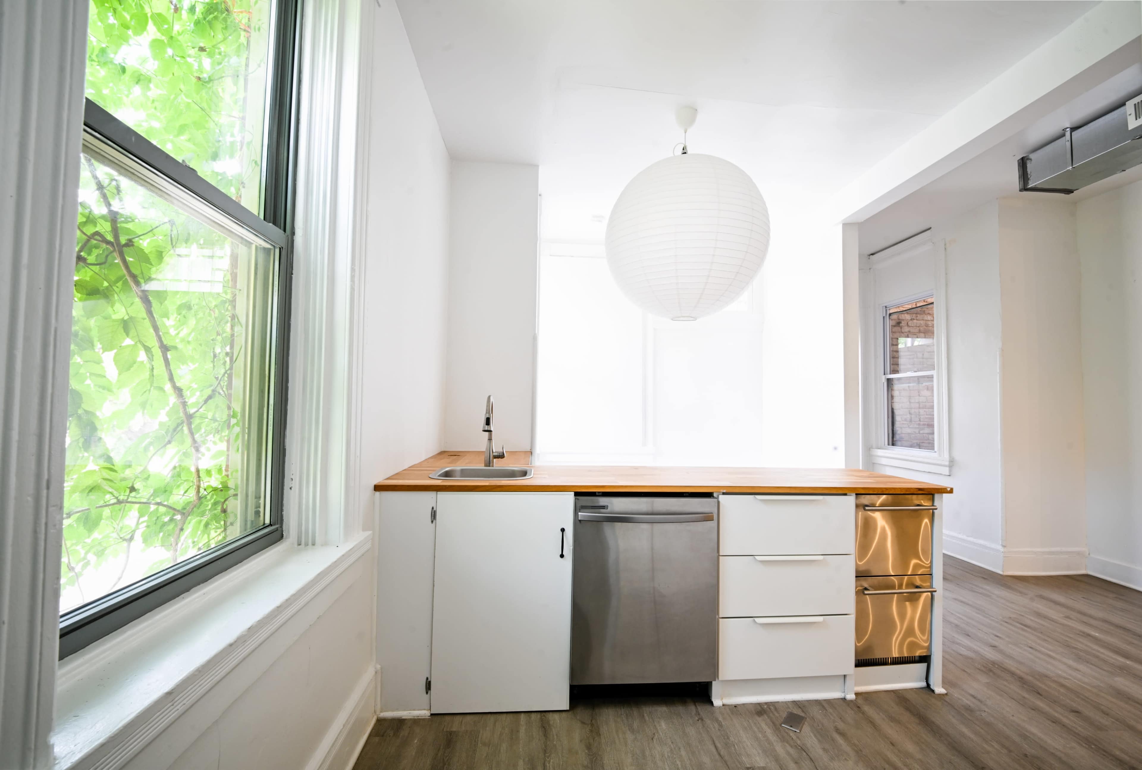 The image shows a modern kitchen with a wooden countertop, stainless steel dishwasher, and large windows allowing natural light to enter the space.
