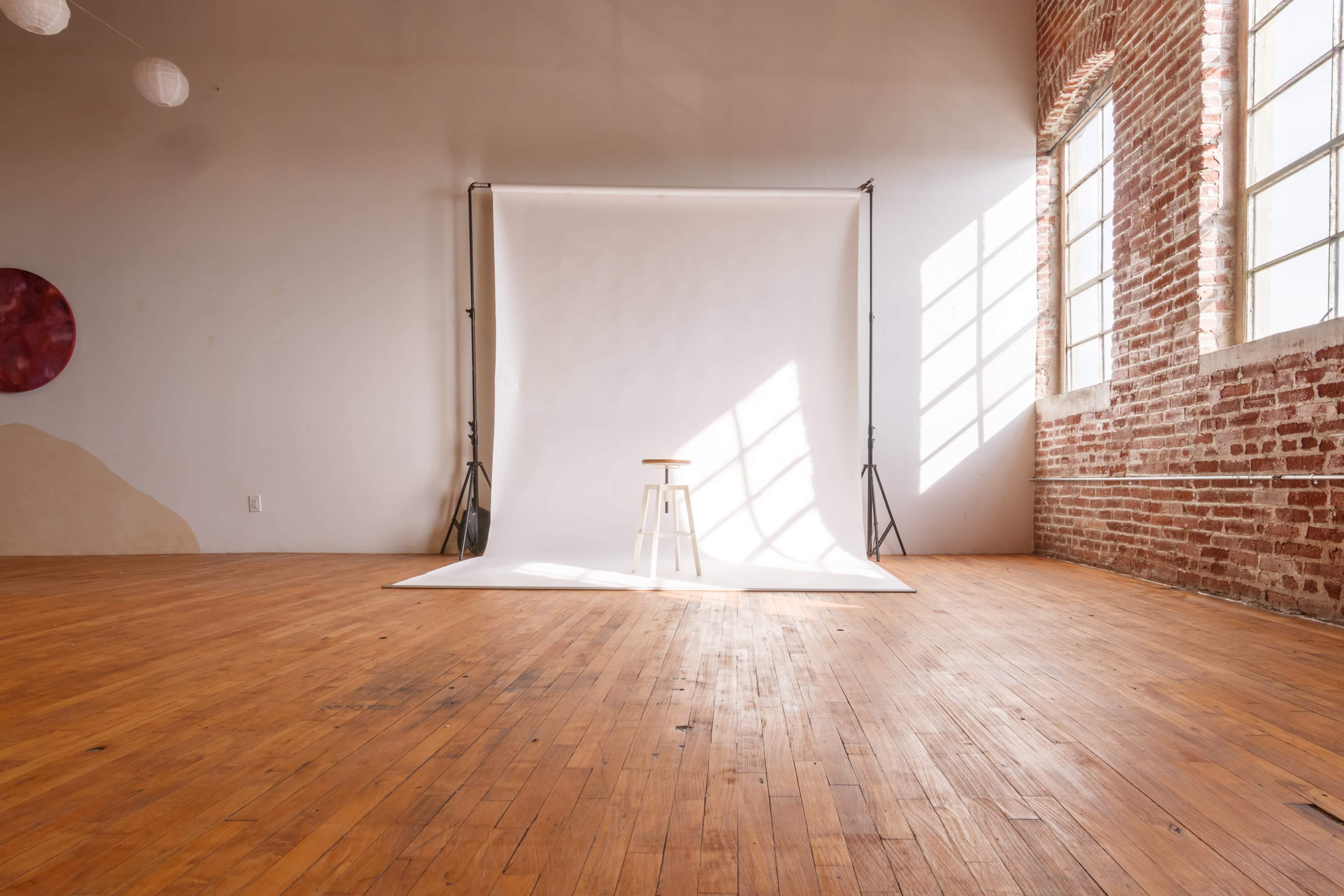 A wooden stool is positioned on a white backdrop in a spacious room with exposed brick walls and large windows allowing natural light to stream in.