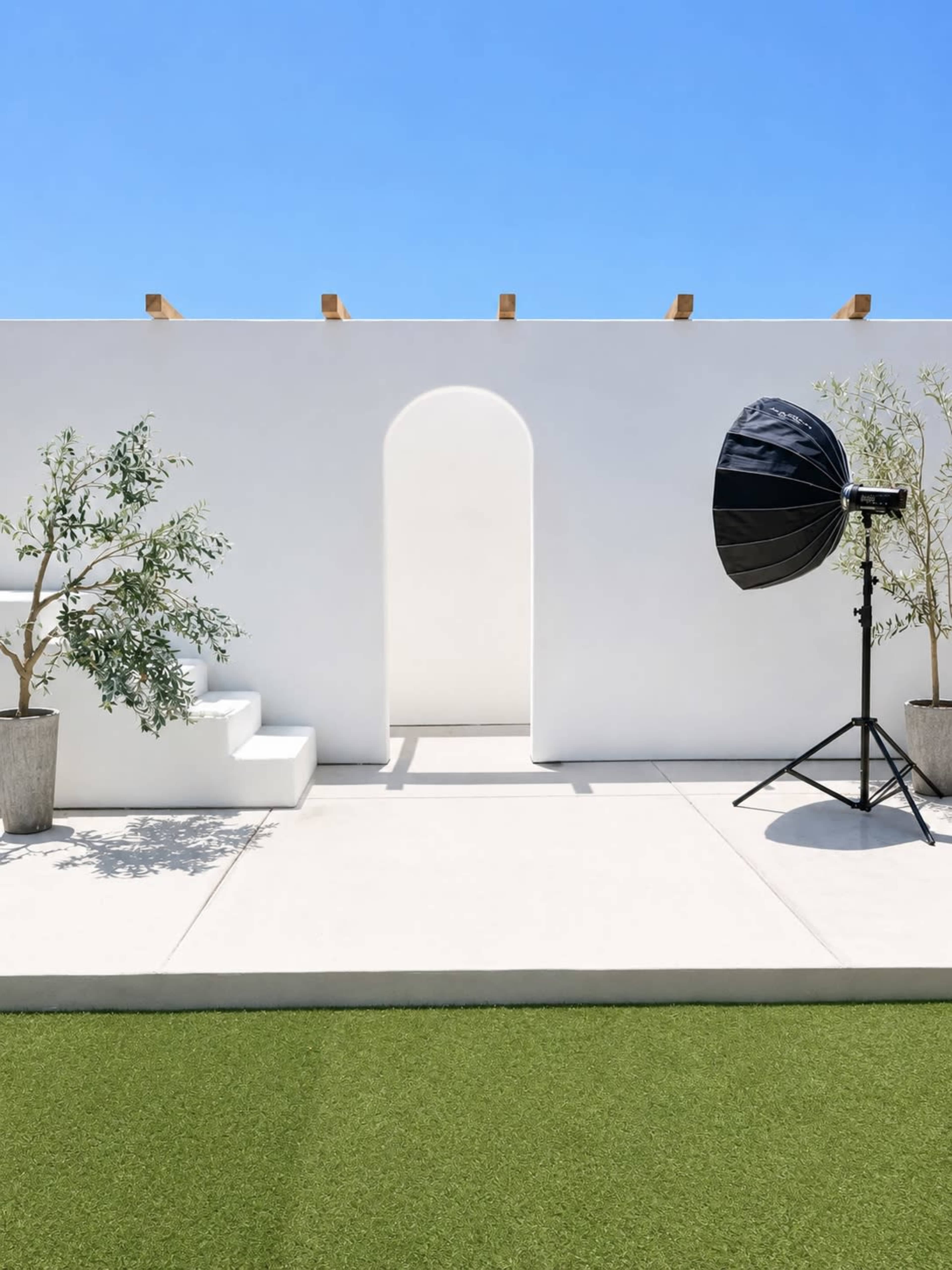 The image shows a modern outdoor space featuring a white wall with an archway, two potted olive trees, steps, and a photography light stand on a green lawn.