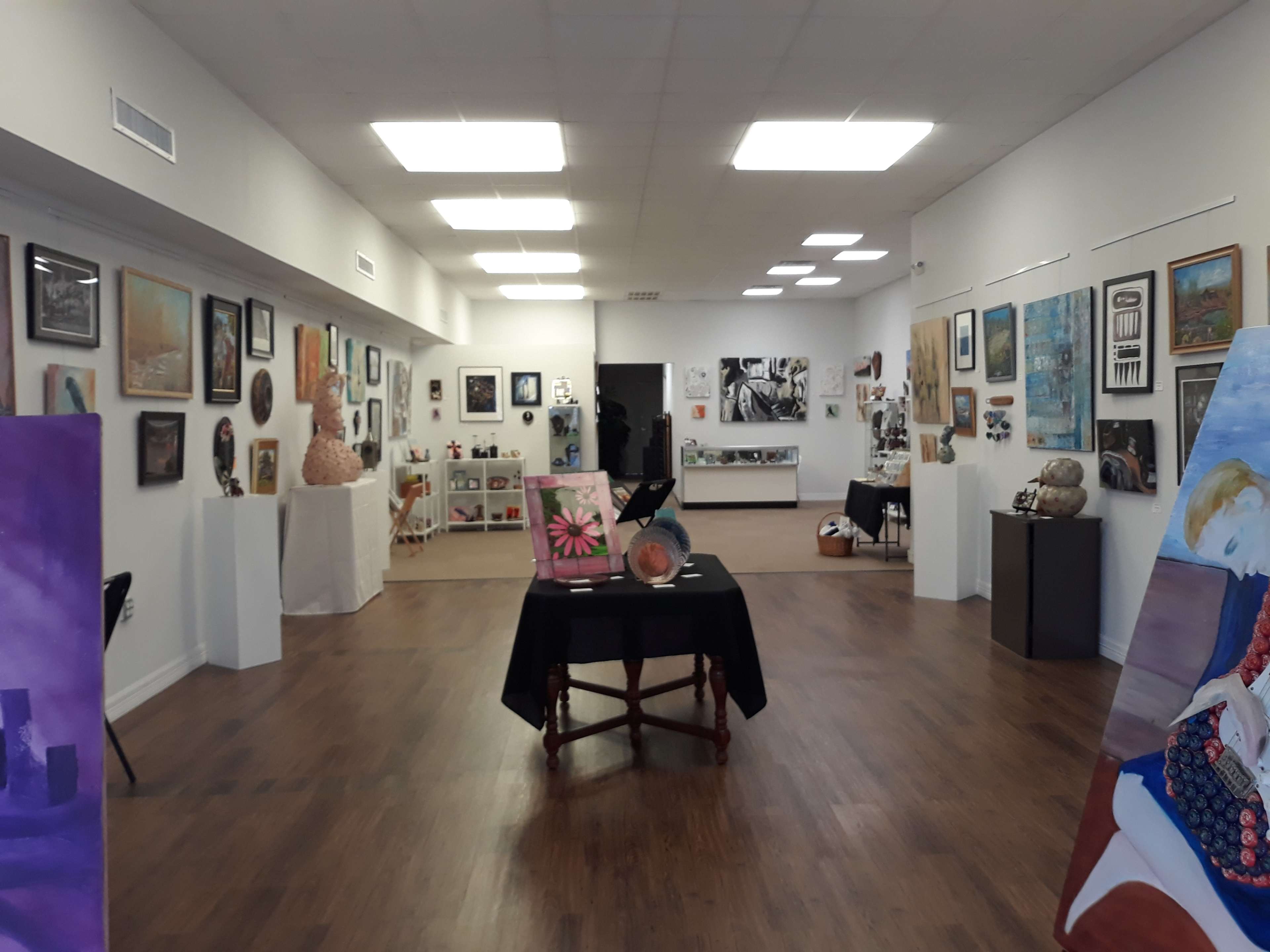 An art gallery features a variety of paintings and sculptures displayed on white walls with hardwood flooring.