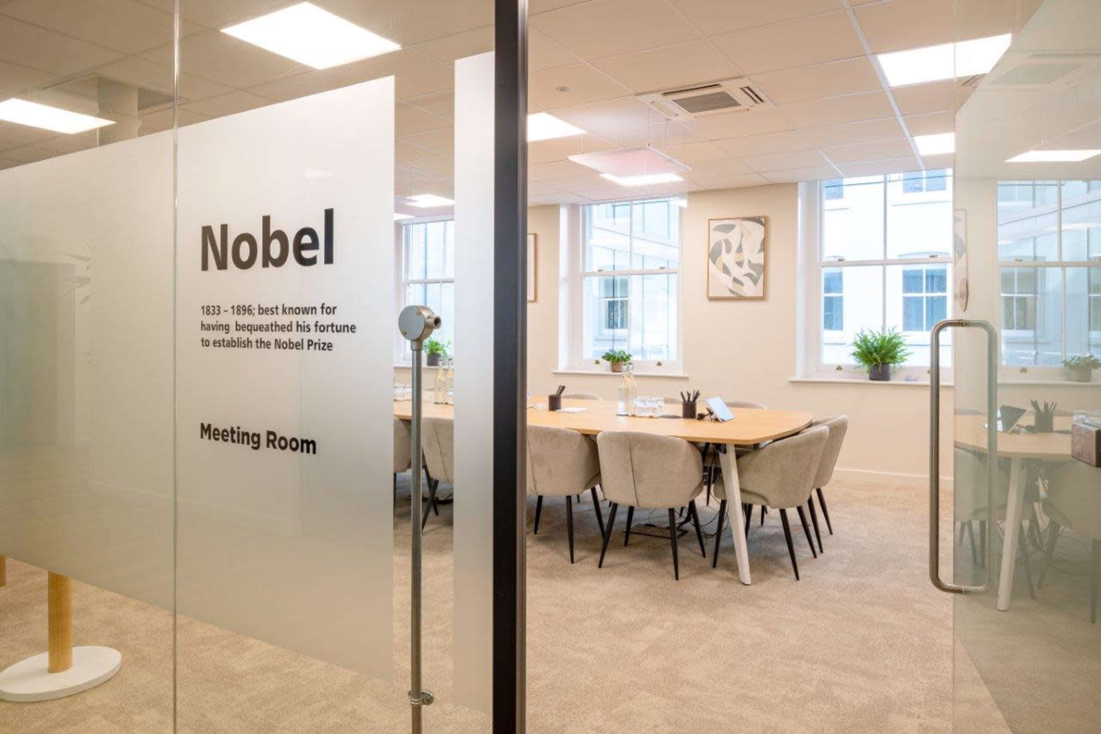 The image shows a modern meeting room labeled "Nobel," featuring a large table surrounded by upholstered chairs and plants near the windows.